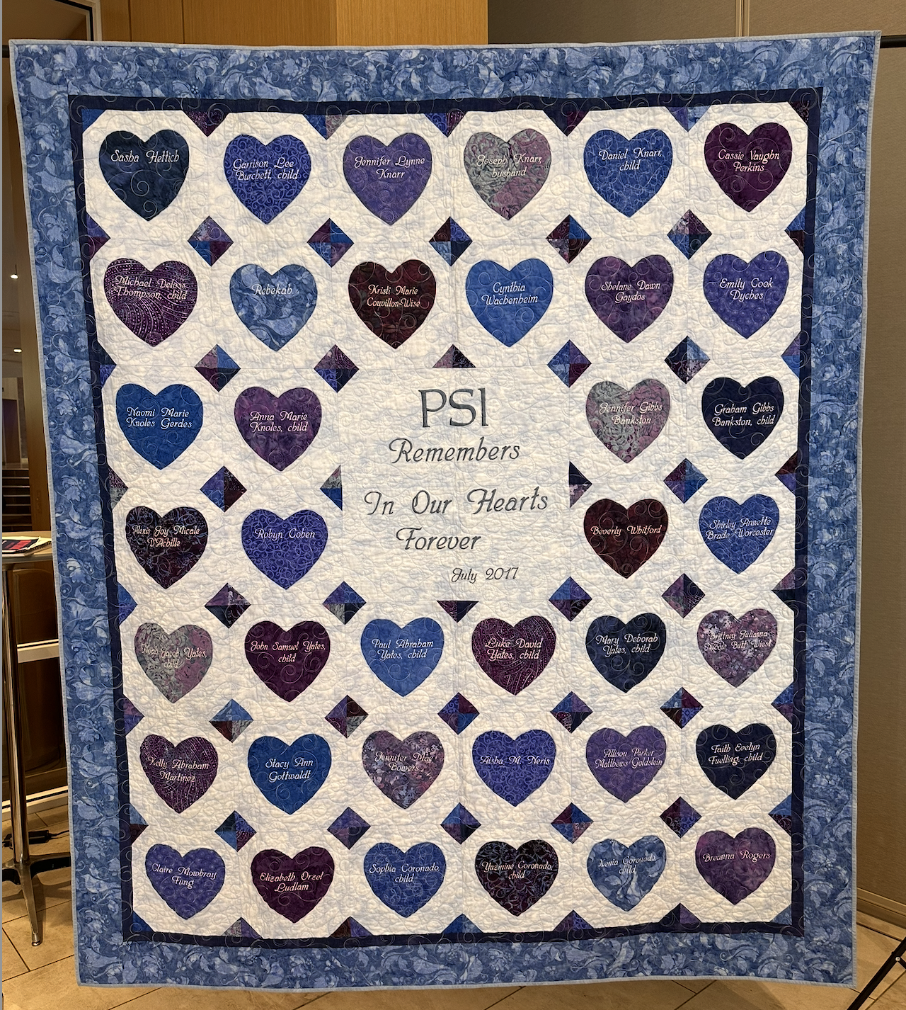 A quilt with a central message that reads 'PSI Remembers In Our Hearts Forever July 2017.' The quilt features rows of hearts in shades of purple, blue, and maroon, each with a child's or individual's name embroidered inside, surrounded by a blue floral border.