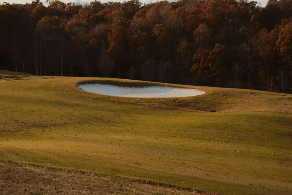 A golf course with a sand trap and water hazard, surrounded by trees with autumn foliage.