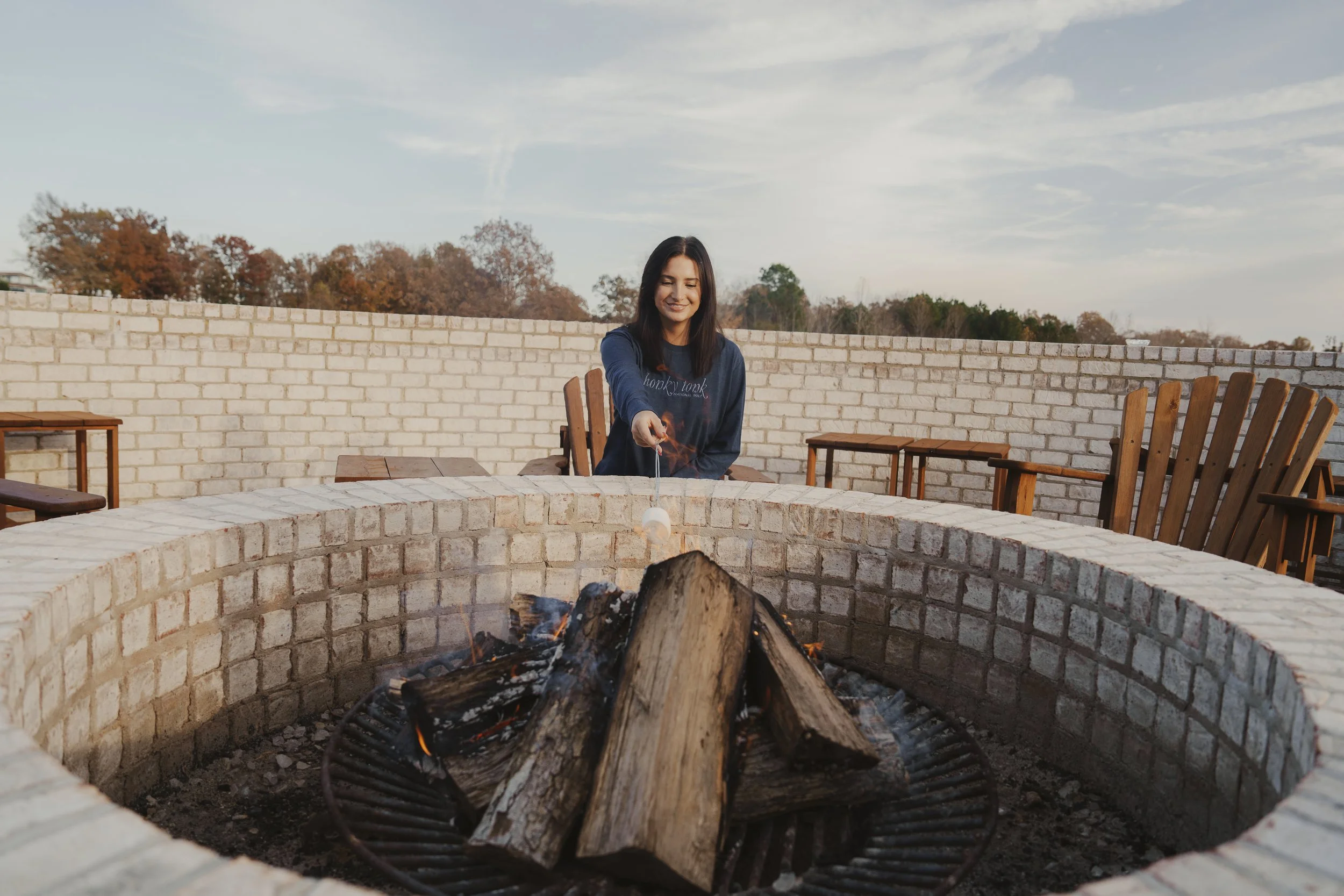 A woman is roasting marshmallows over a fire pit on a brick patio during daytime.