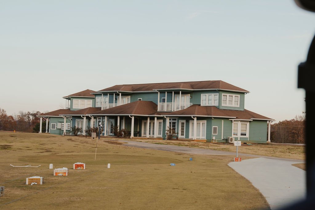 A large, two-story house with light blue siding, white trim, and a brown roof, situated on a spacious lawn with a dirt driveway.