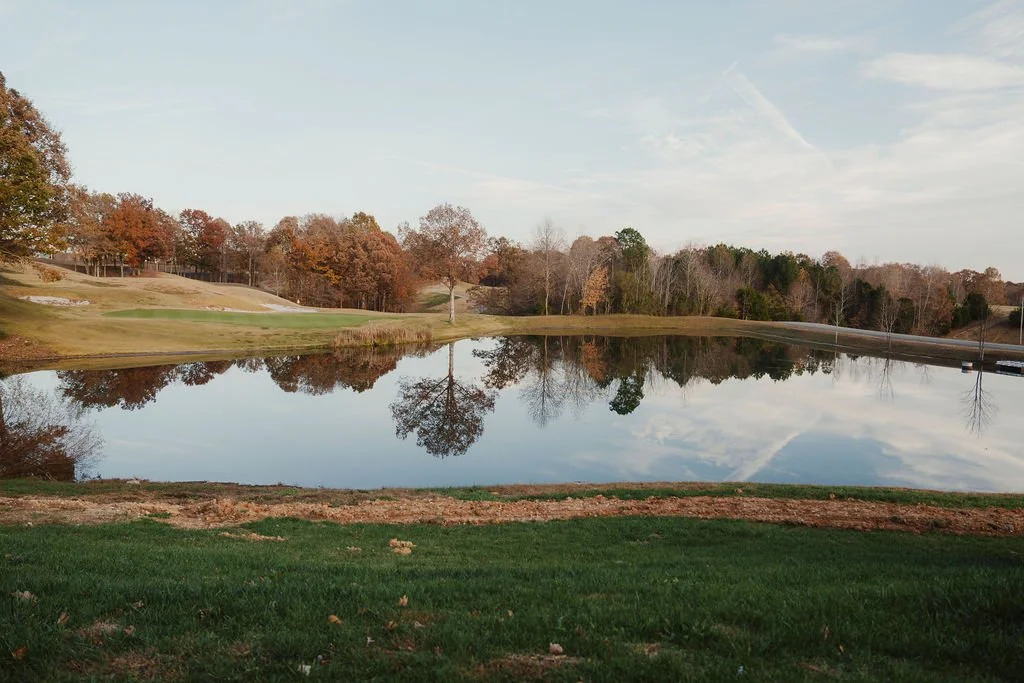 A peaceful golf course with a pond reflecting trees and sky, during autumn.