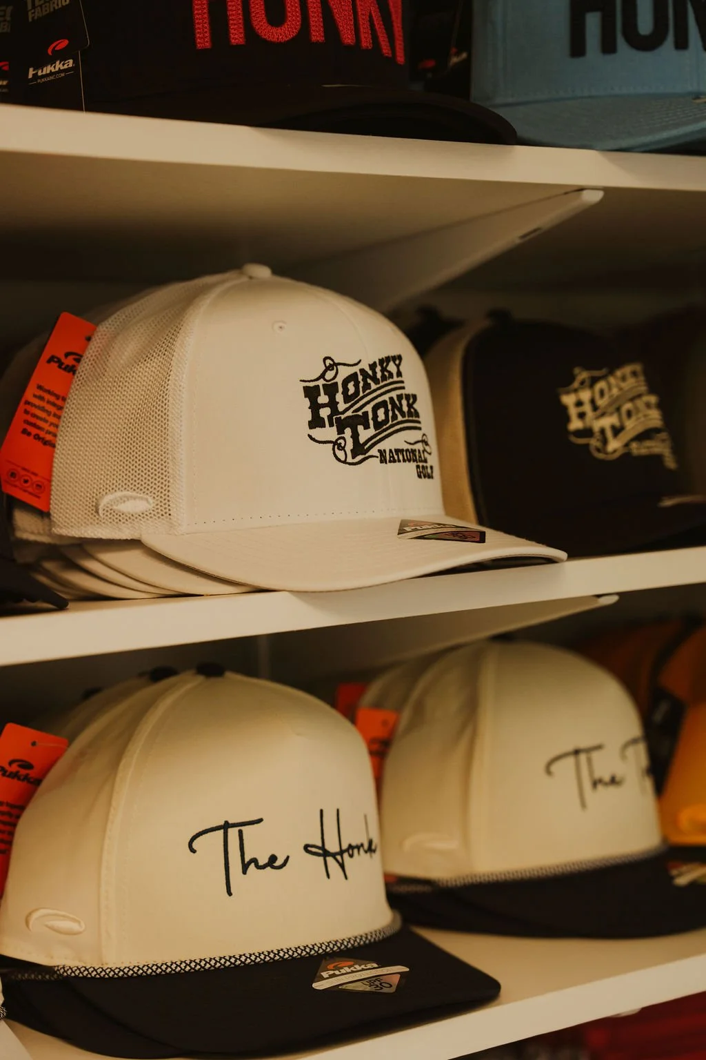 White baseball cap with black text reading 'The Honky Tonk' on a store shelf, with other caps in the background.