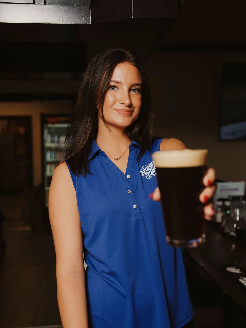 A young woman with dark hair wearing a blue sleeveless shirt, standing in a bar, holding a glass of dark beer and smiling at the camera.
