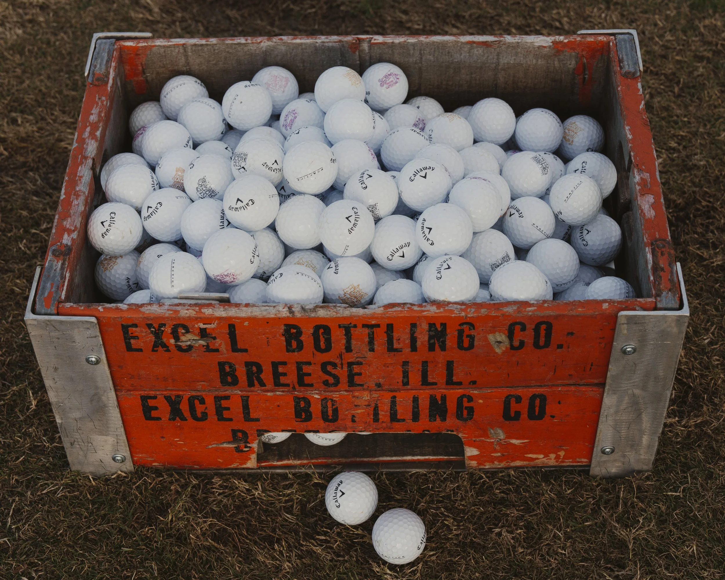 A wooden crate filled with many white golf balls, with some balls spilled on the grass outside the crate.