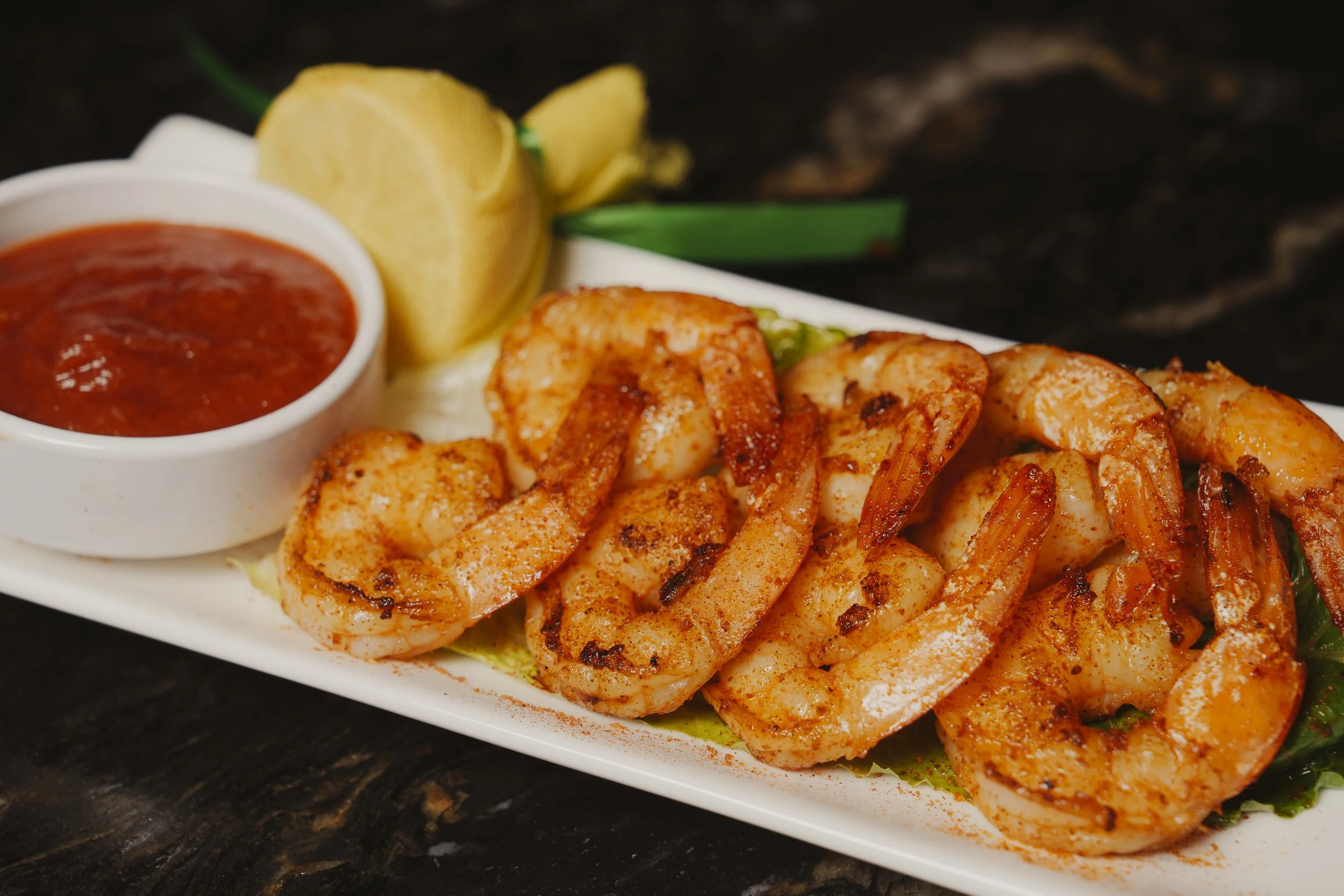 Grilled shrimp with a side of red sauce, lemon slices, and green garnish on a white plate.