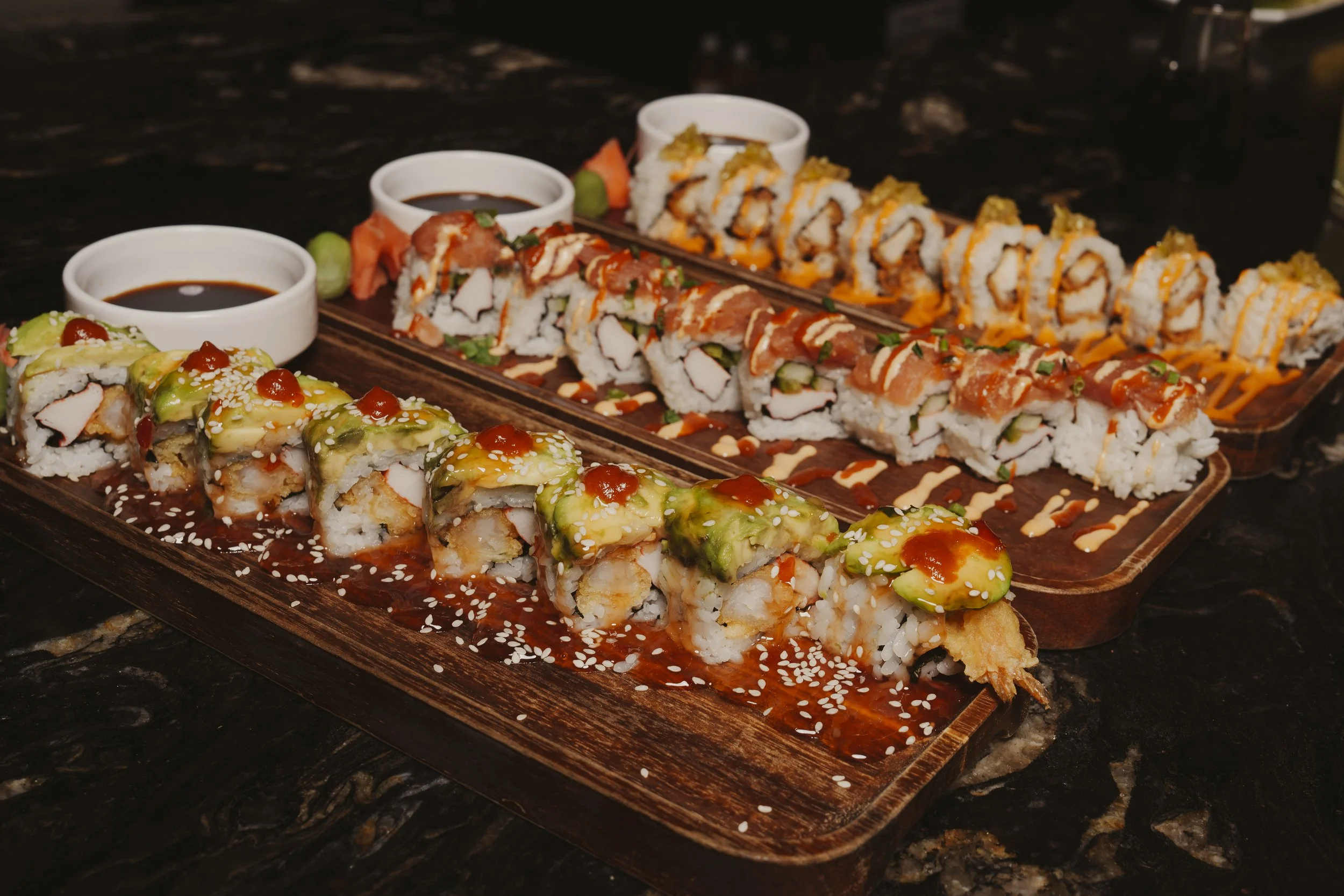 Two wooden sushi platters with assorted sushi rolls, garnished with sauces, sesame seeds, and small bowls of soy sauce, on a dark marble surface.