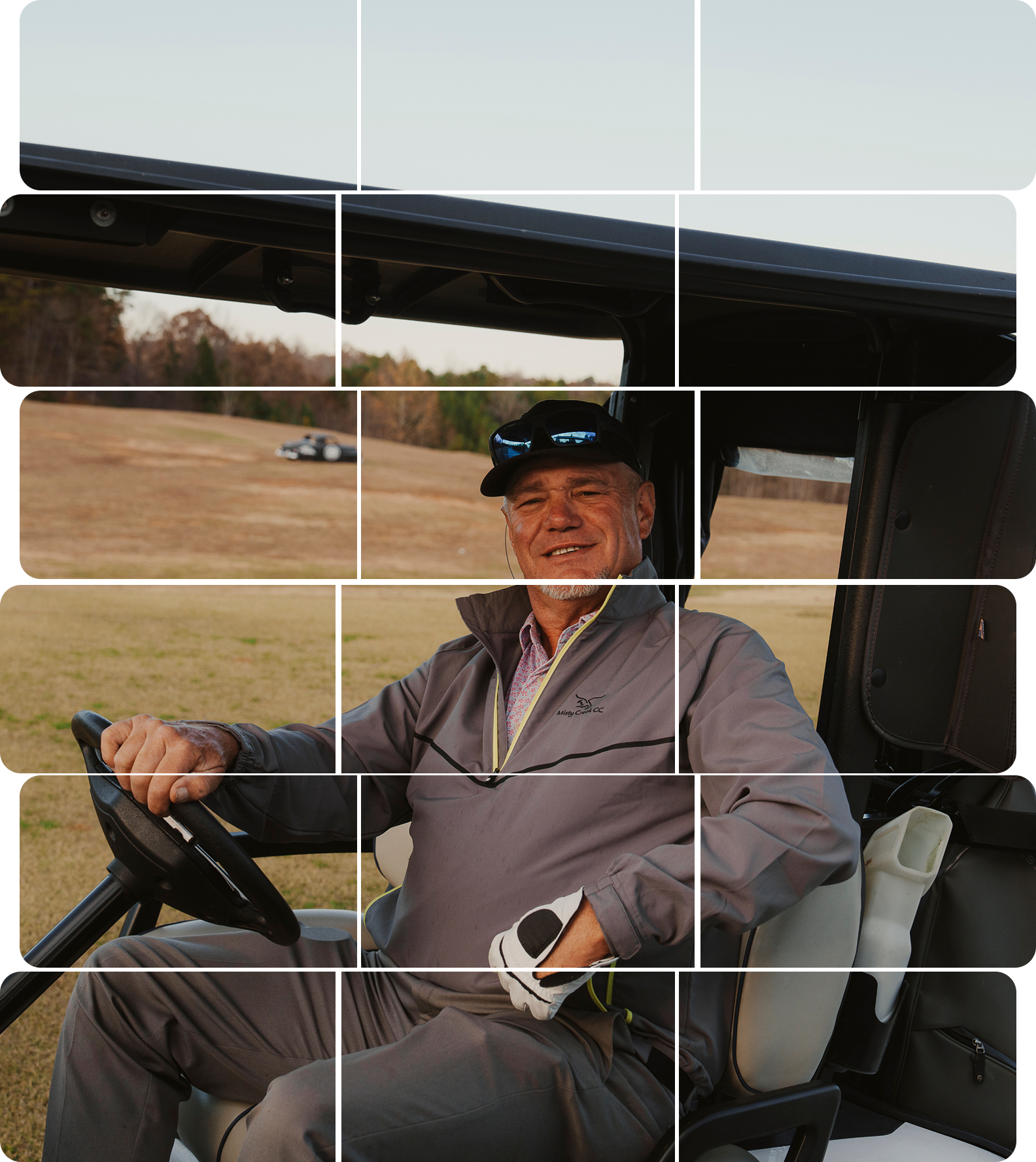 A man smiling and sitting in a golf cart on a golf course, wearing a cap, sunglasses, a gray jacket, and golf gloves.
