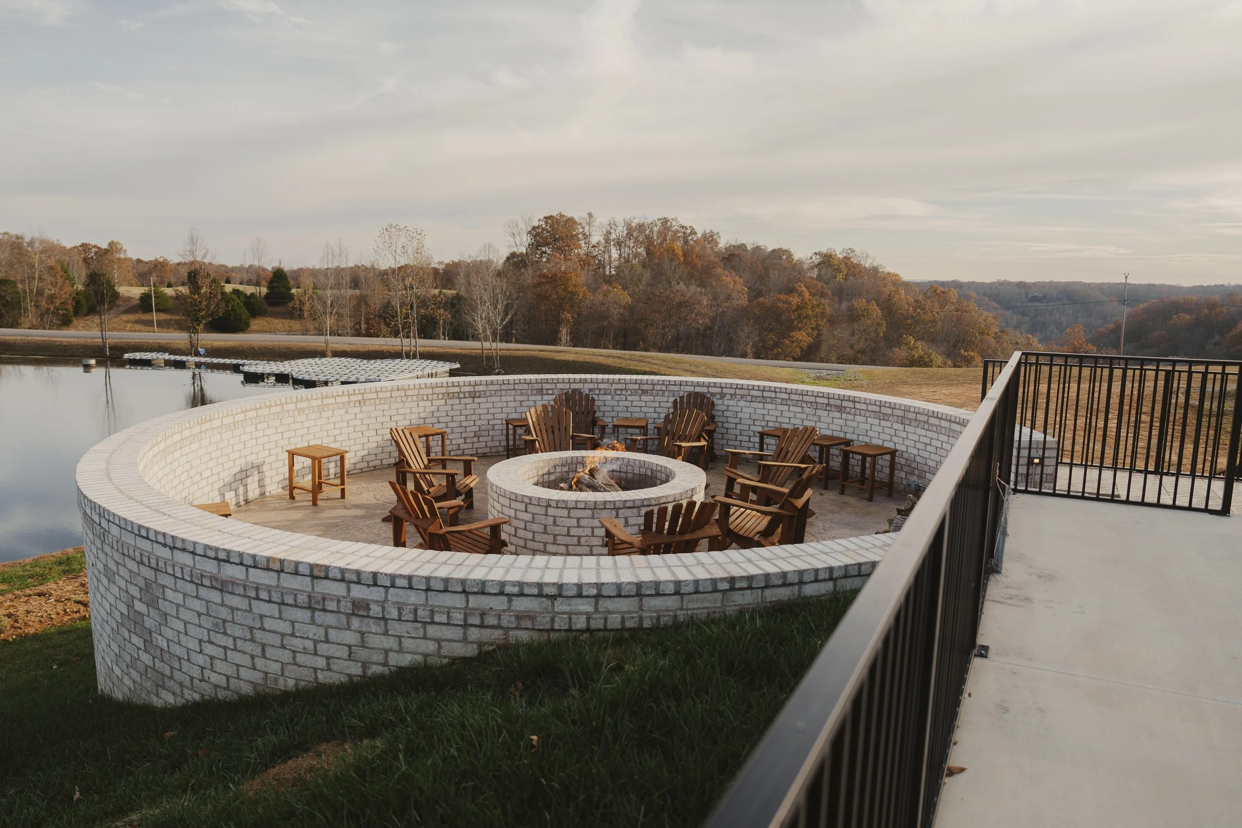 Outdoor seating area with a fire pit surrounded by wooden chairs, enclosed by a curved white brick wall, overlooking a lake and a landscape with trees and cloudy sky in the background.