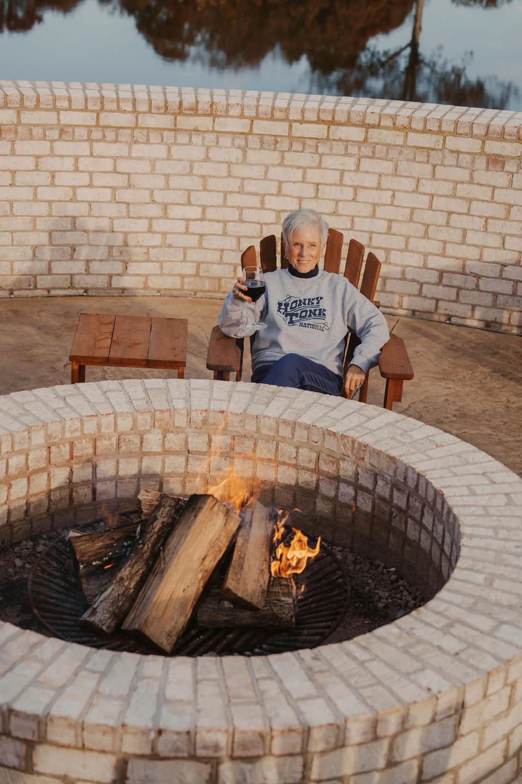 An elderly woman with gray hair sitting in an Adirondack chair, holding a glass of red wine, on a patio with a brick fire pit and a small wooden table, near a body of water with trees reflected on its surface.