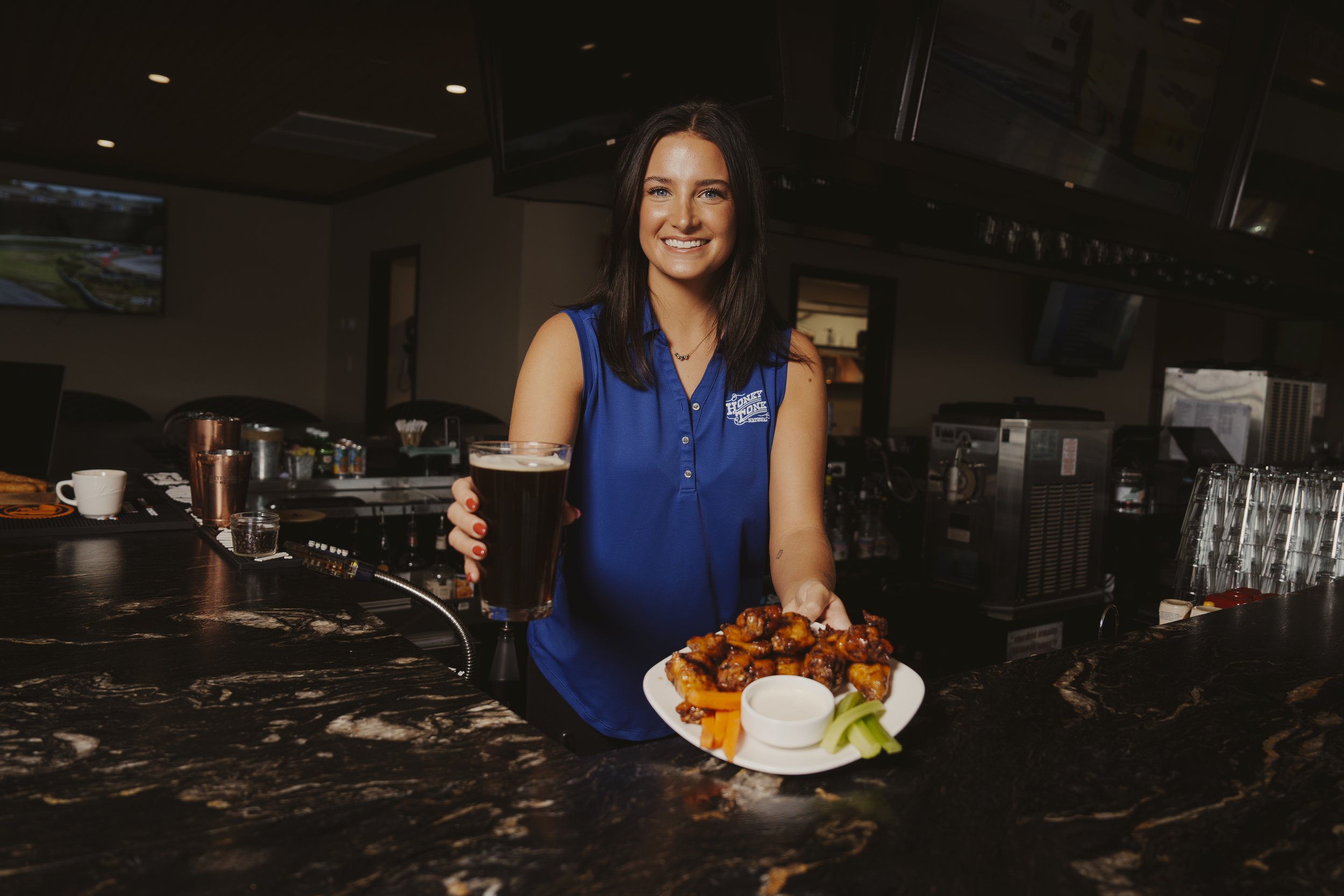 A woman in a blue sleeveless shirt smiles and holds a pint of dark beer in one hand while presenting a plate of wings, celery, carrots, and dipping sauce in a bar or restaurant setting.