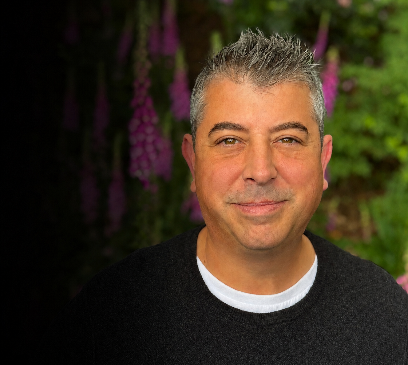 A smiling man with gray hair and brown eyes, wearing a black sweater and a white shirt underneath, standing outdoors with a blurred background of green foliage and purple flowers.