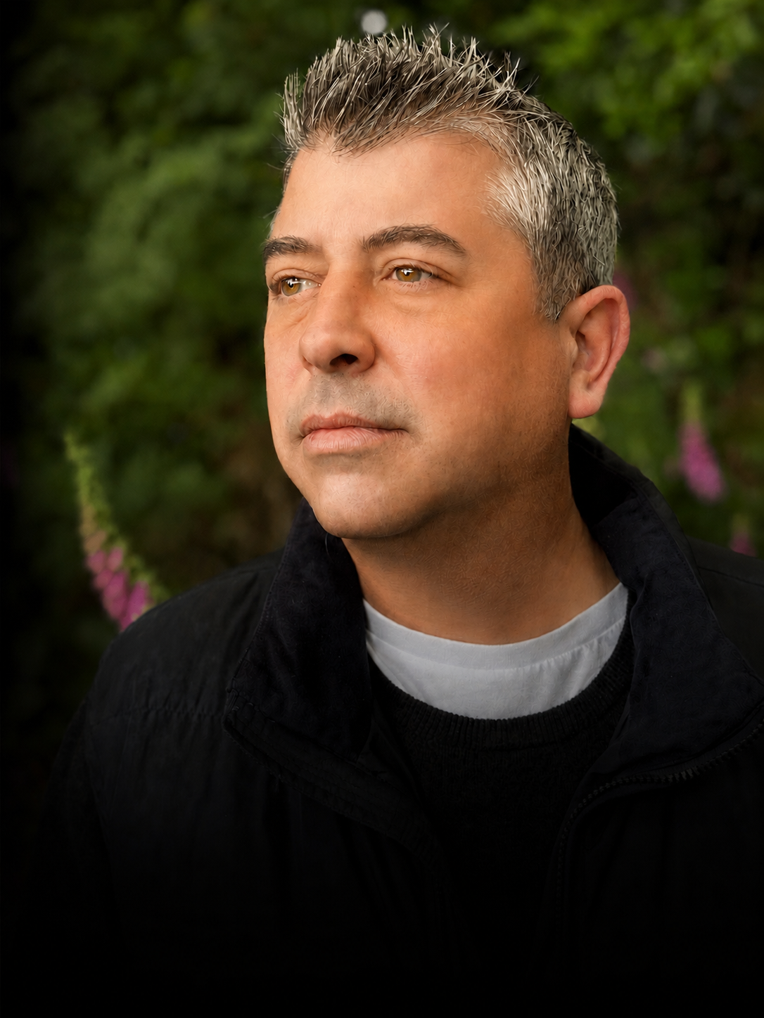 A man with short, spiked gray hair and light skin gazes into the distance, outdoors with green foliage and purple flowers in the background.