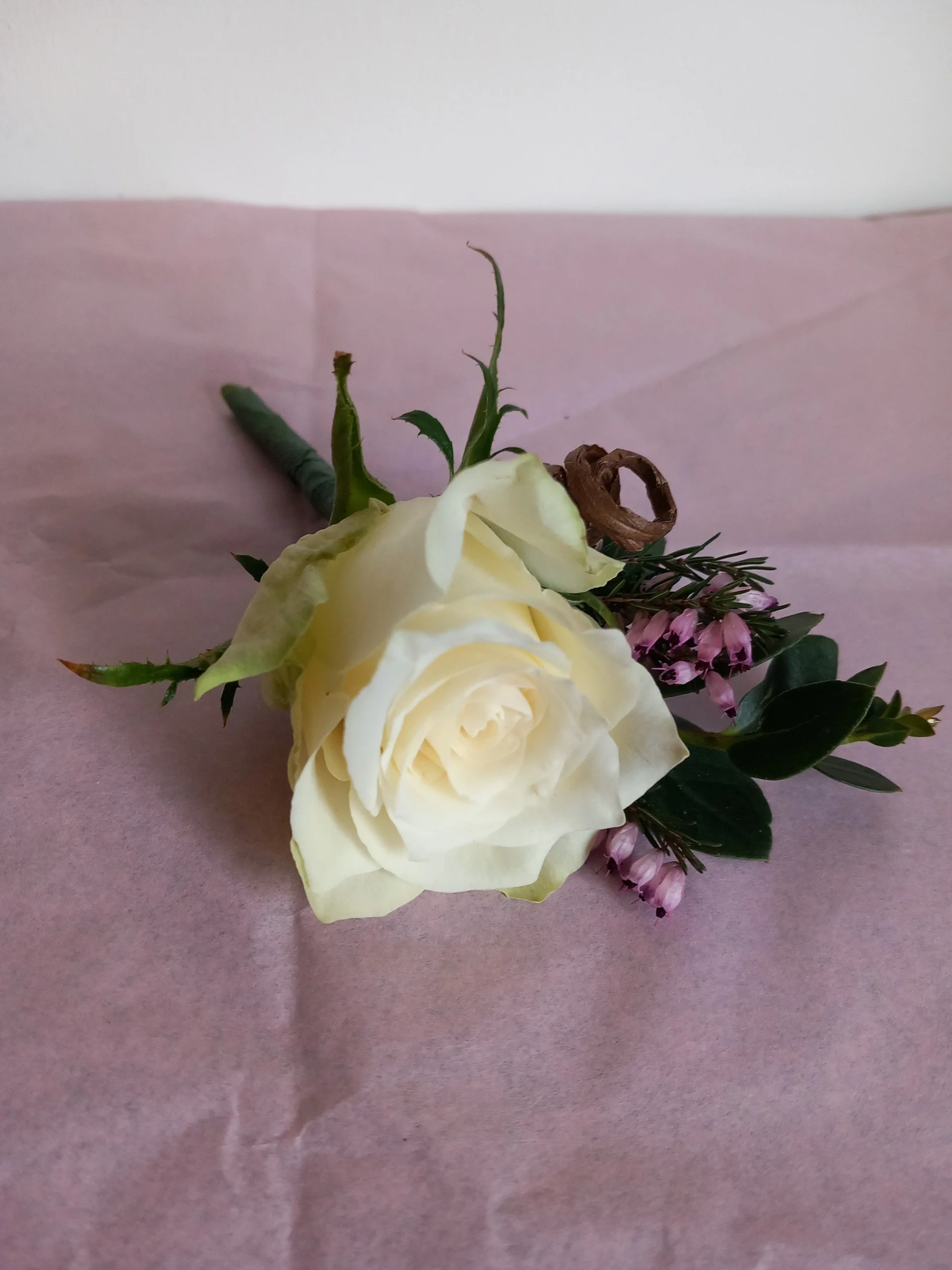White rose buttonhole with greenery and small purple heather with a brown bohemian ribbon.