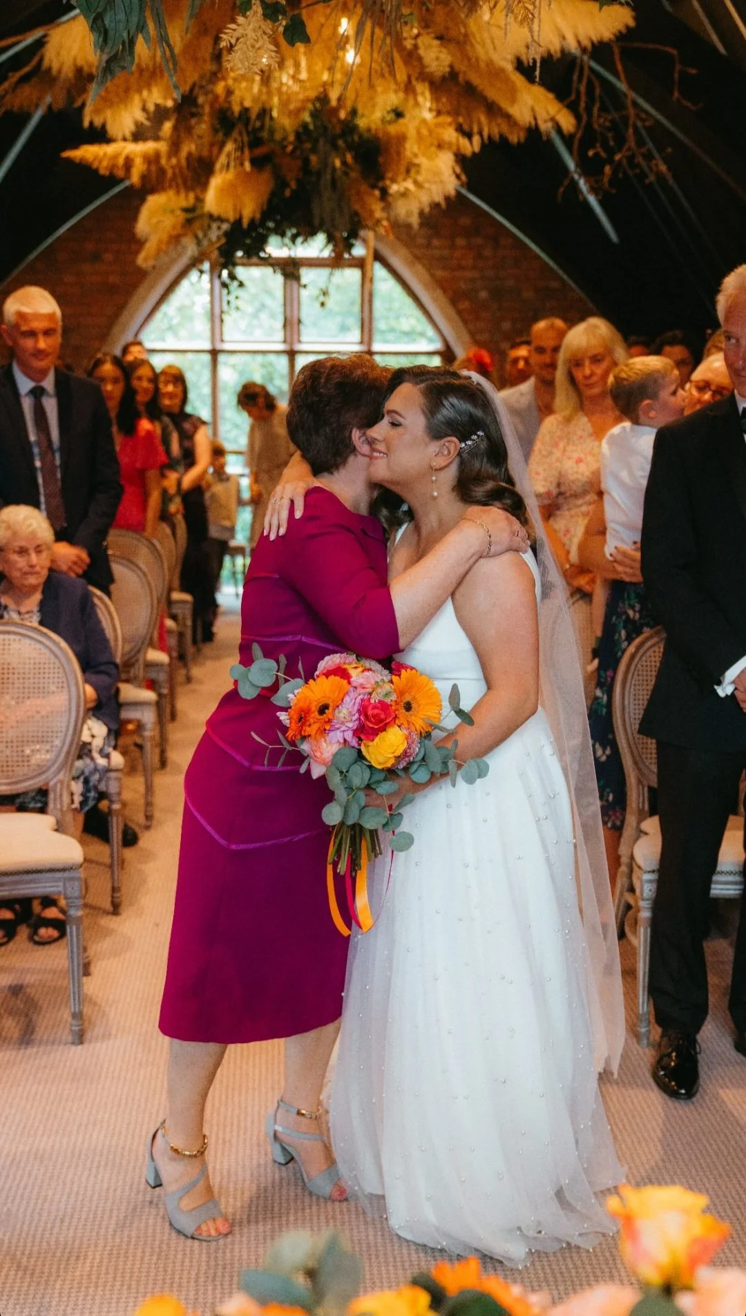 A bride in a white wedding dress hugging a woman in a magenta dress, who is holding a colorful bouquet of flowers, at a wedding ceremony with guests in the background.