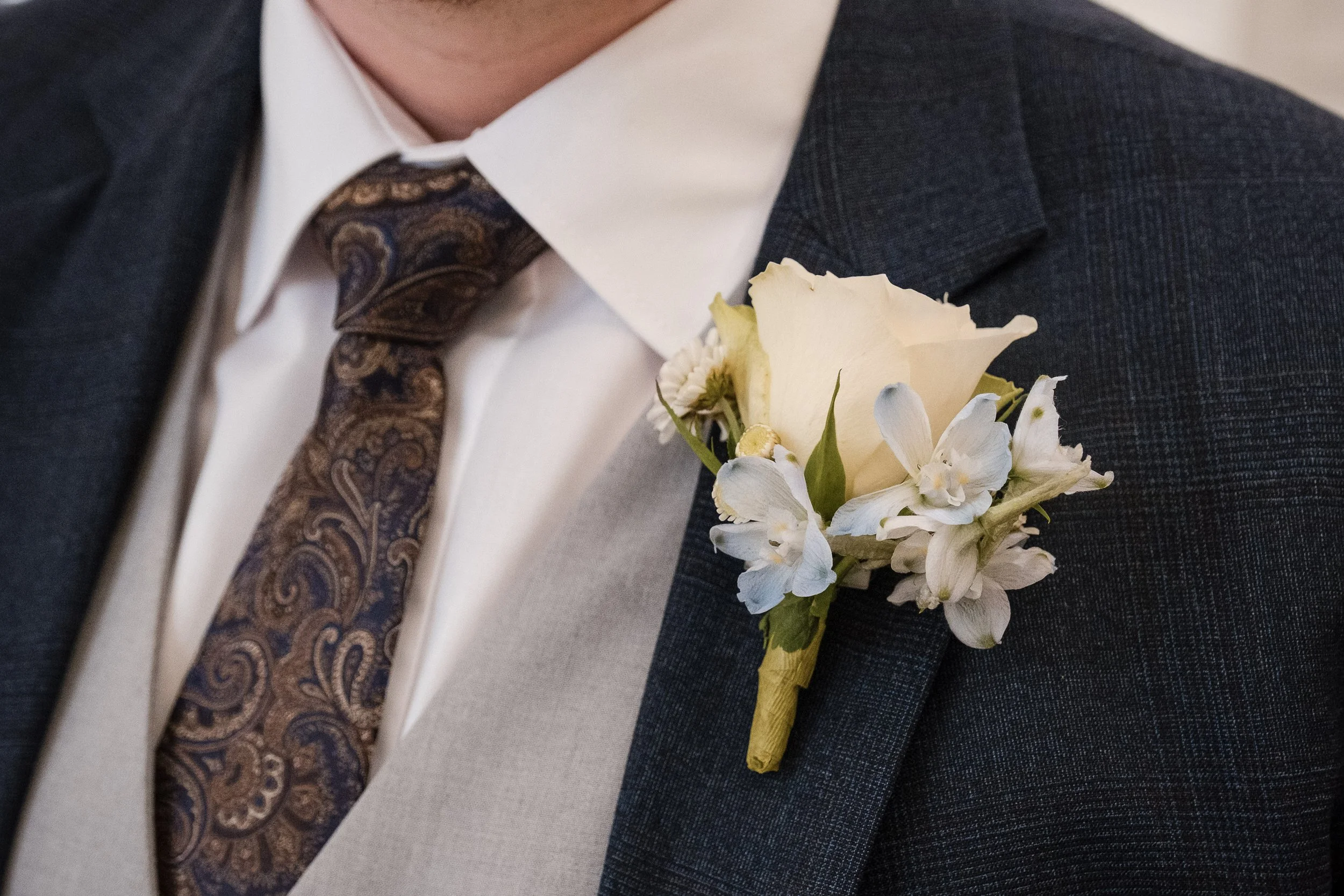 A man wearing a dark suit, white shirt, and paisley tie with a white flower boutonniere on his lapel.