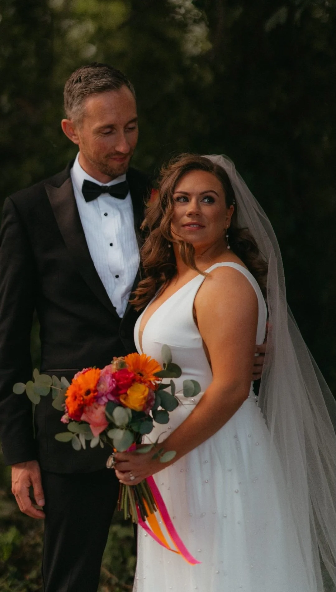 A bride and groom pose together outdoors during their wedding, with the bride holding a colorful bouquet and the groom wearing a black tuxedo.