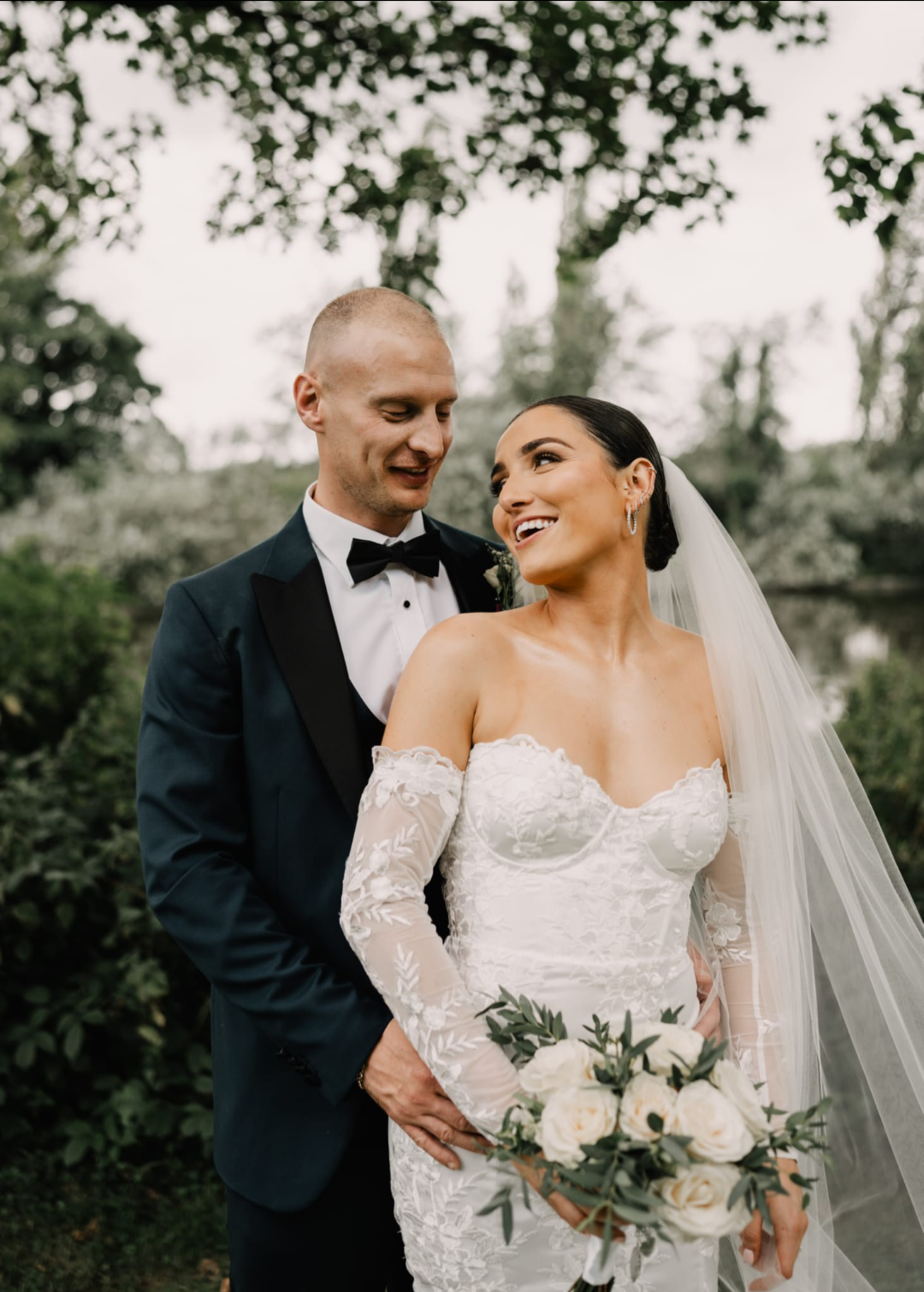 A newlywed couple smiling outdoors, the bride in a lace wedding gown and veil holding a bouquet of white roses, the groom in a black tuxedo with bow tie, surrounded by greenery.