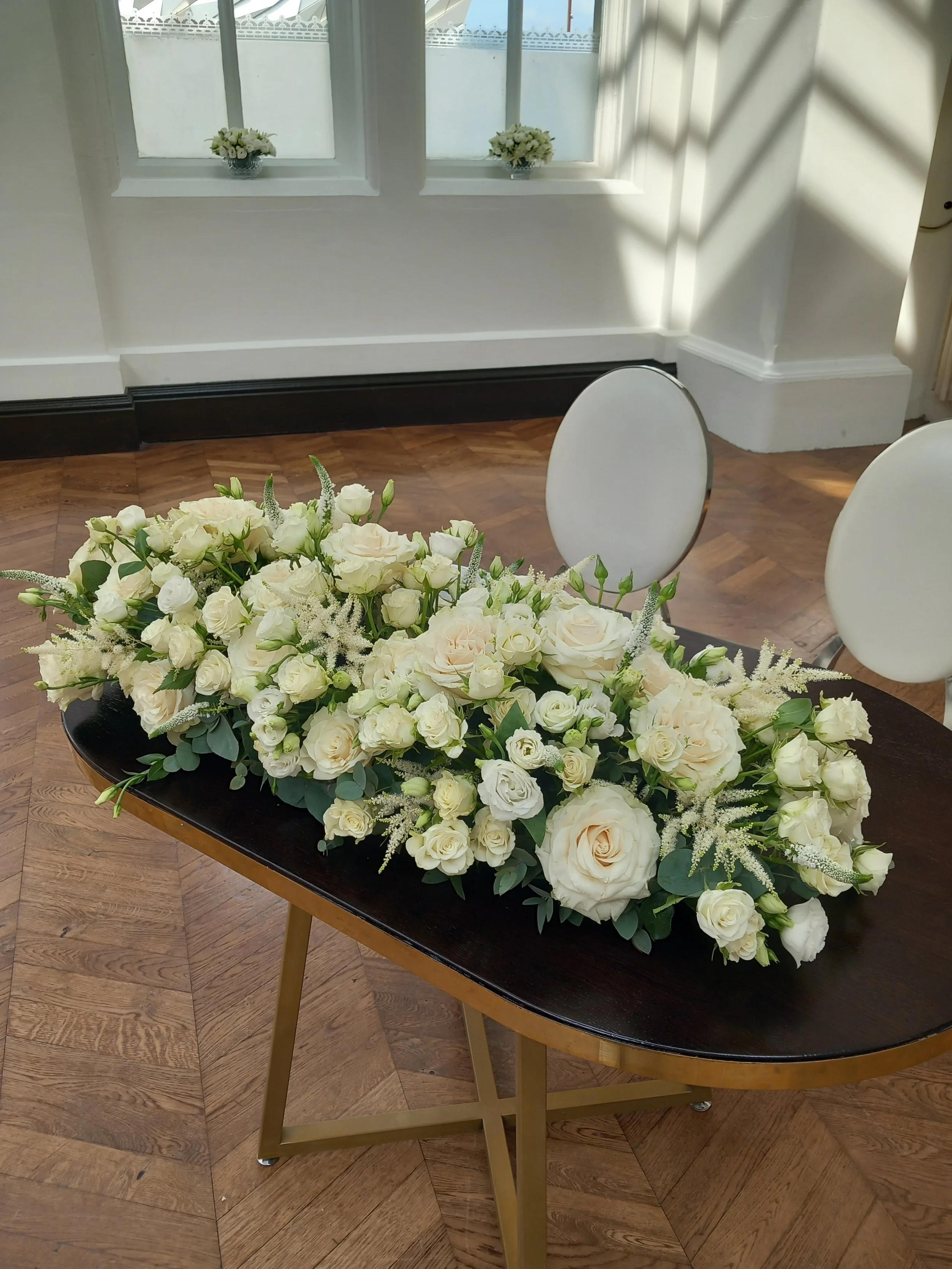 A wedding signing table floral arrangement with white and cream-coloured flowers in the Titanic Hotel Belfast, with white chairs and large windows in the background.