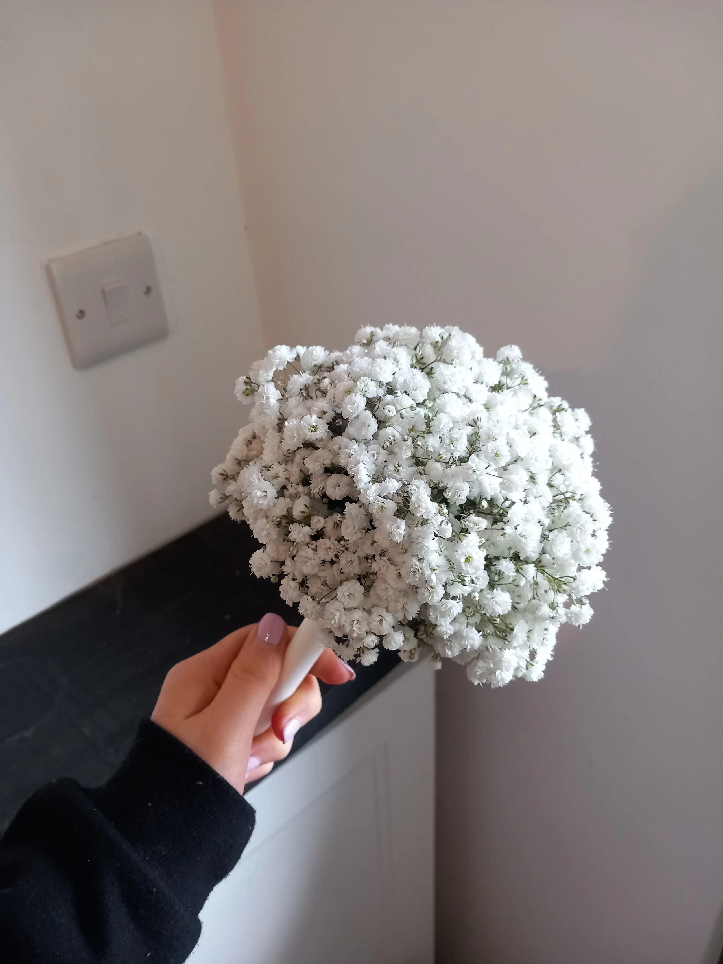A hand holding a small bouquet of white baby's breath flowers in front of a neutral wall and light switch.
