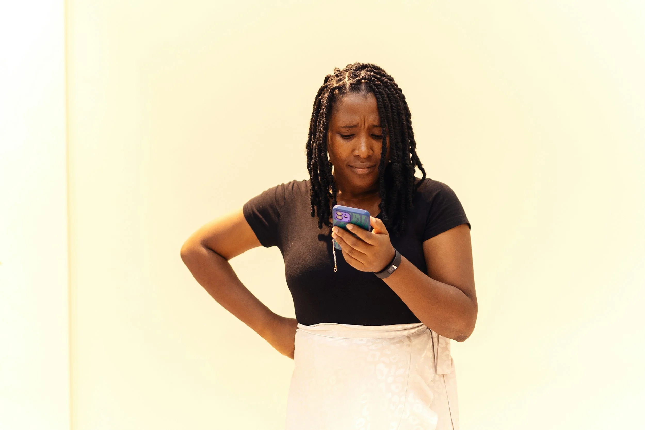 A woman frowns in confusion while looking at her smartphone, standing with her hand on her hip against a plain light background.