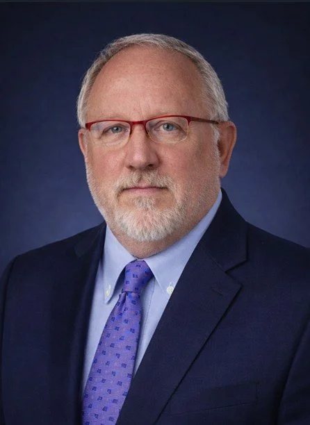 A middle-aged man with light skin, a beard, and glasses with red frames, wearing a dark suit, light blue shirt, and purple tie, posed against a dark blue background.