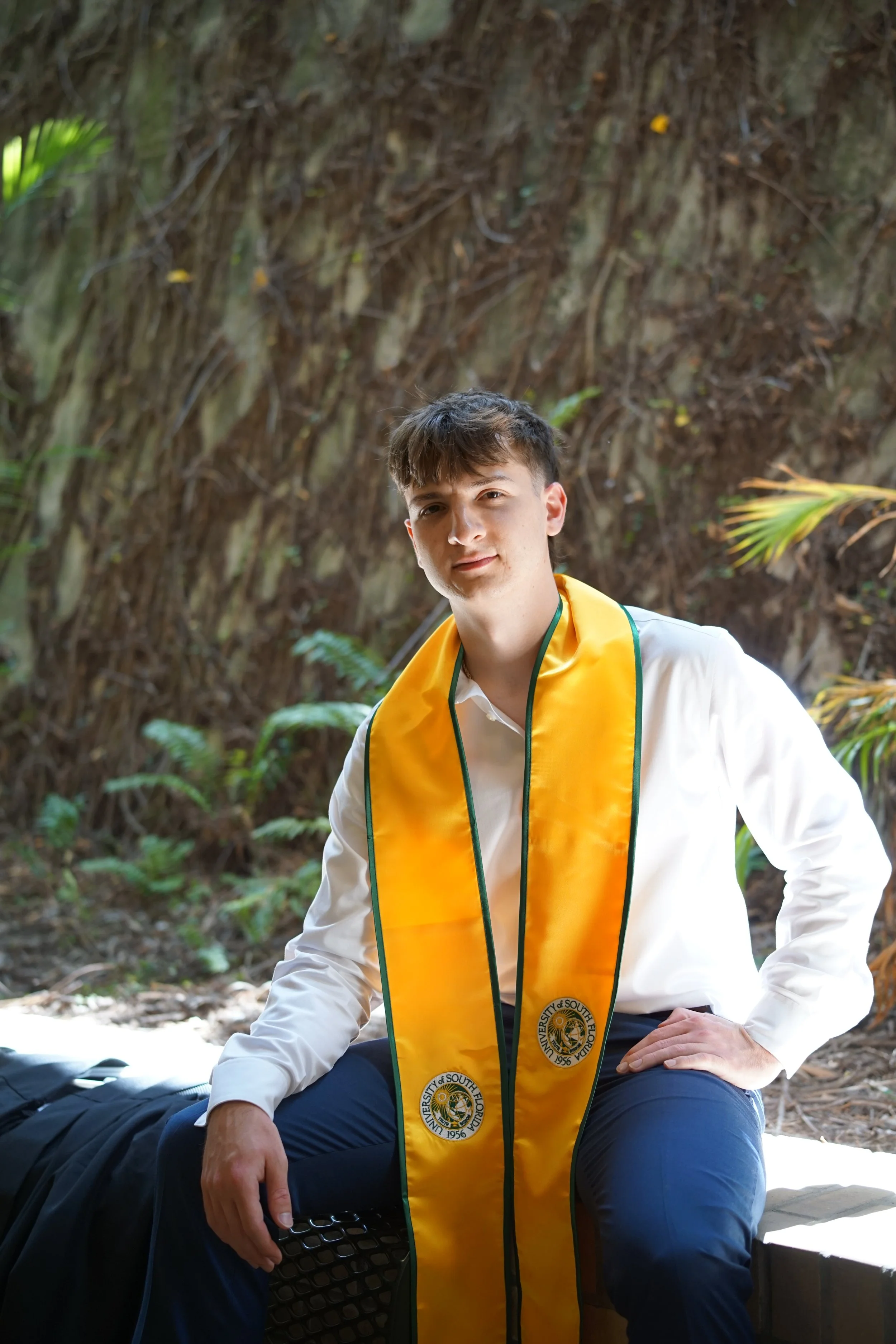 A young man with brown hair wearing a white shirt and a yellow and green graduation stole, sitting outdoors in a wooded area with greenery and trees in the background.