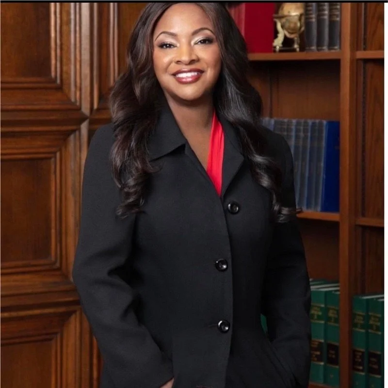 A woman with long wavy black hair wearing a black blazer and red blouse, standing in front of a wooden bookshelf with books.
