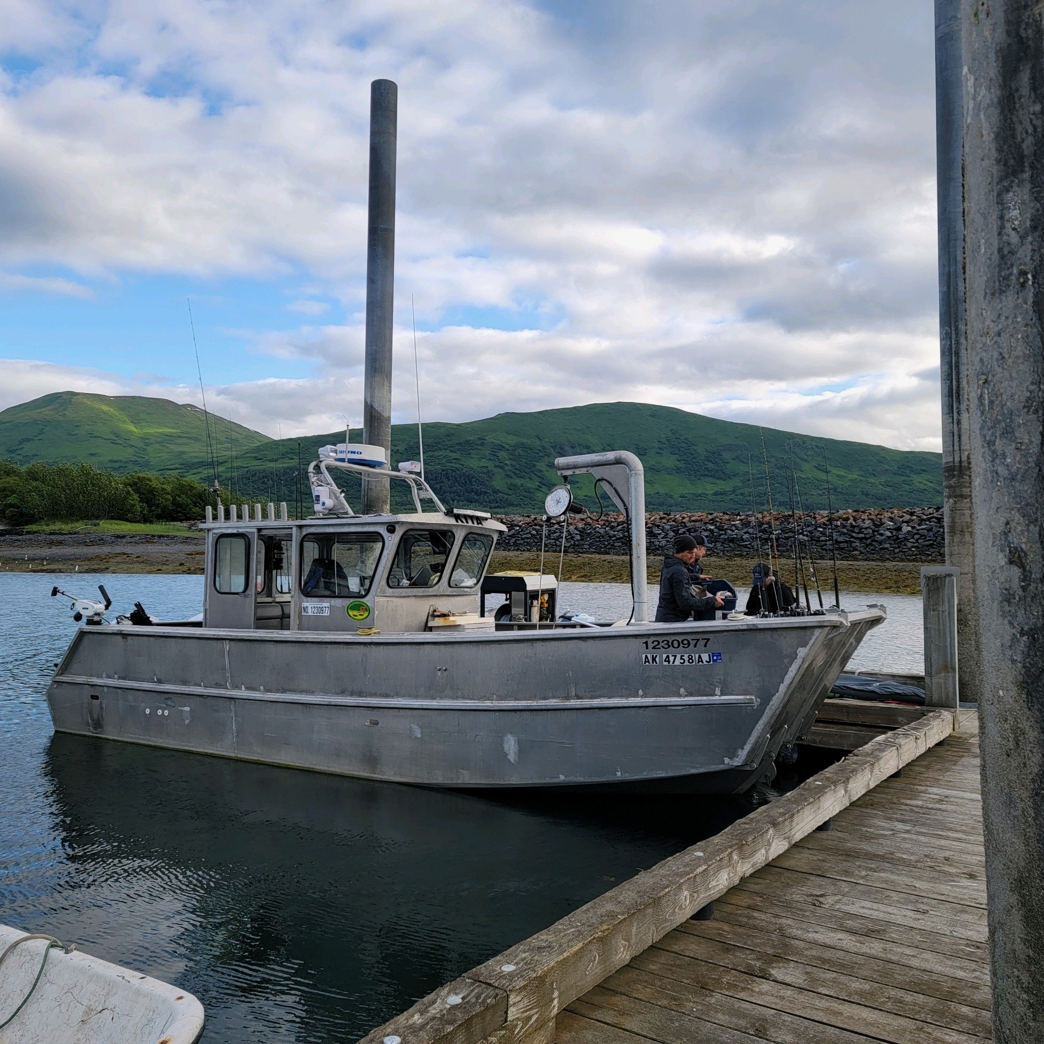 Fishing boat docked at a pier with a person on board, mountains and trees in the background, under a partly cloudy sky.