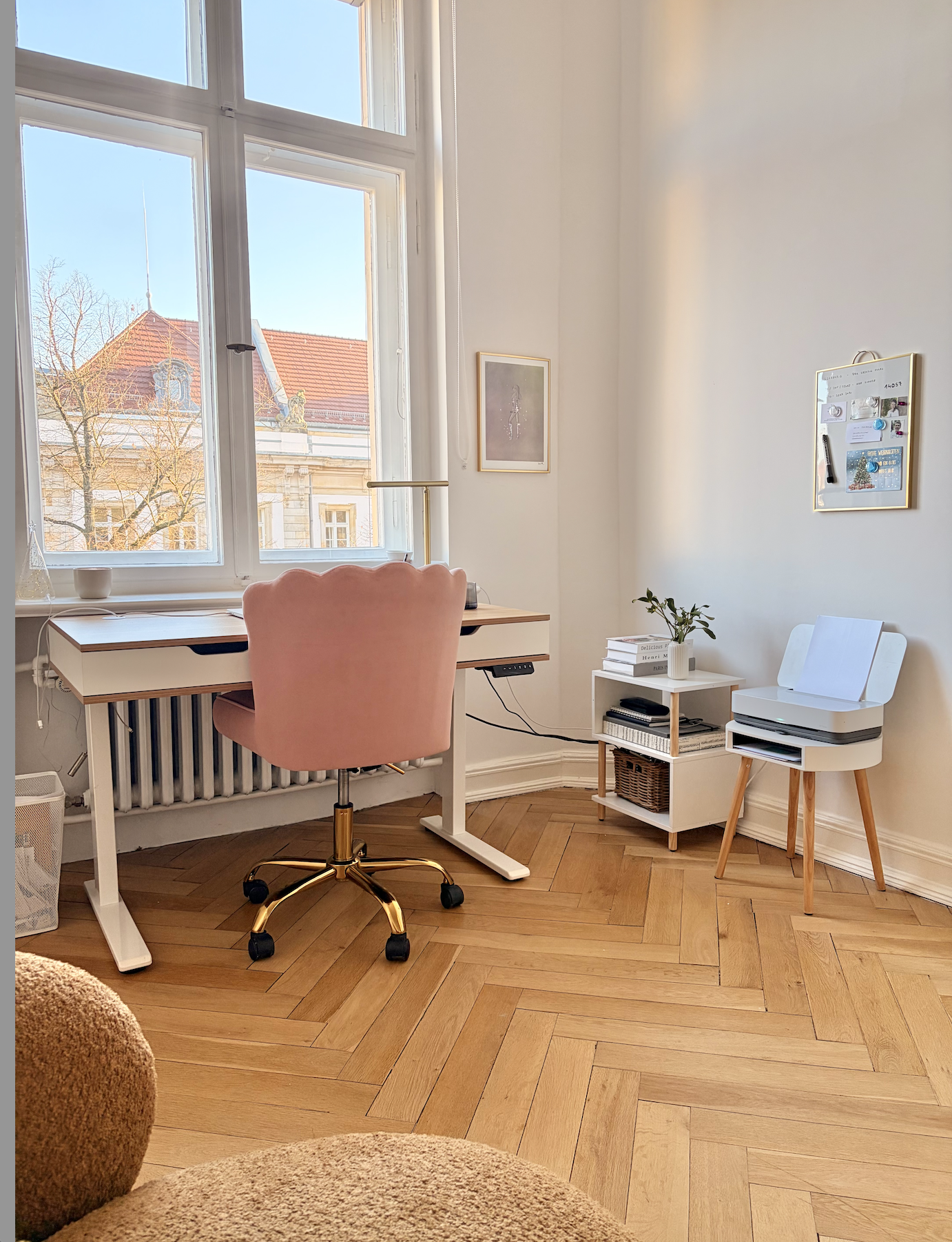 Ein hell erleuchtetes Arbeitszimmer von Christiane Germann mit einem großen Fenster, einem Schreibtisch mit rosa Bürostuhl, einem kleinen weißen Regal mit Pflanzen und Büchern, und einem Drucker auf einem kleinen weißen Beistelltisch.
