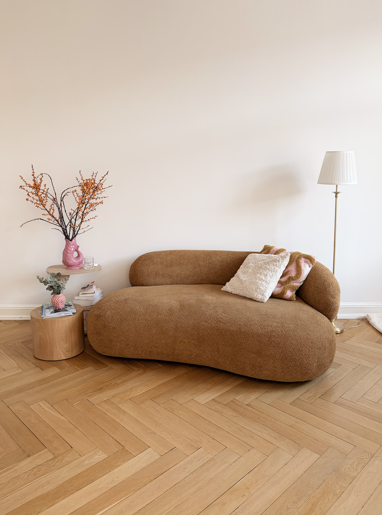 Ein Wohnzimmer von Christiane Germann mit einem braunen, modernen Sofa, einem kleinen beigen Holztablett mit dekorativer Vase, einem Stapel Zeitschriften, einem weißen Kissen und einem link angeordneten pinkfarbenen Vase mit Zweigen.