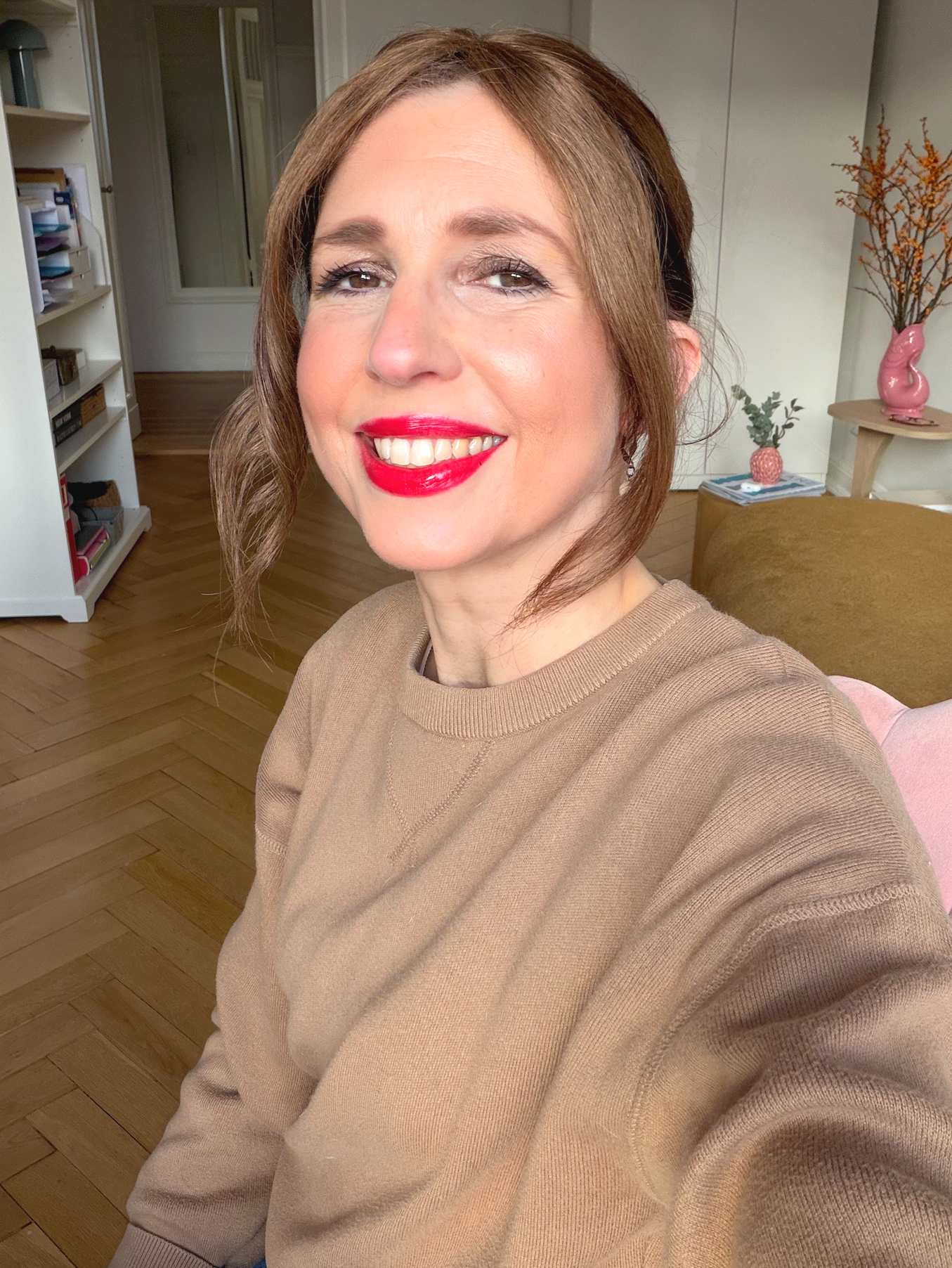 Nahaufnahme von Christiane Germann mit roten Lippen, braunem Haar und beigen Pullover, im Wohnzimmer mit weißen Regalen, Holzfußboden und dekorativen Vasen im Hintergrund.