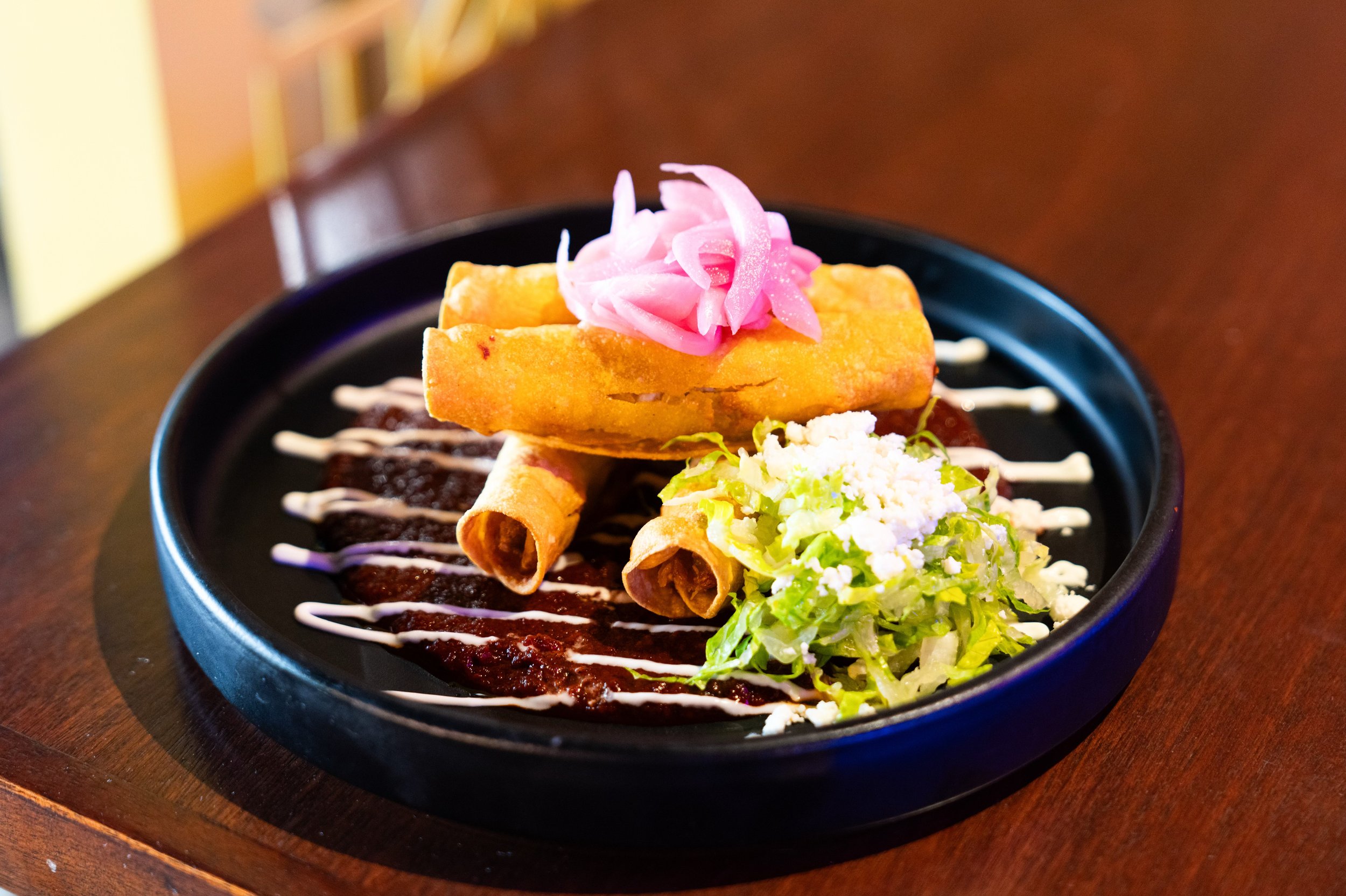 Flautas Bañadas-
Four perfectly crisped chicken flautas, served on a bed of bold, smoky adobo sauce. Topped with vibrant pickled onions, fresh microgreens, and drizzled with crema.