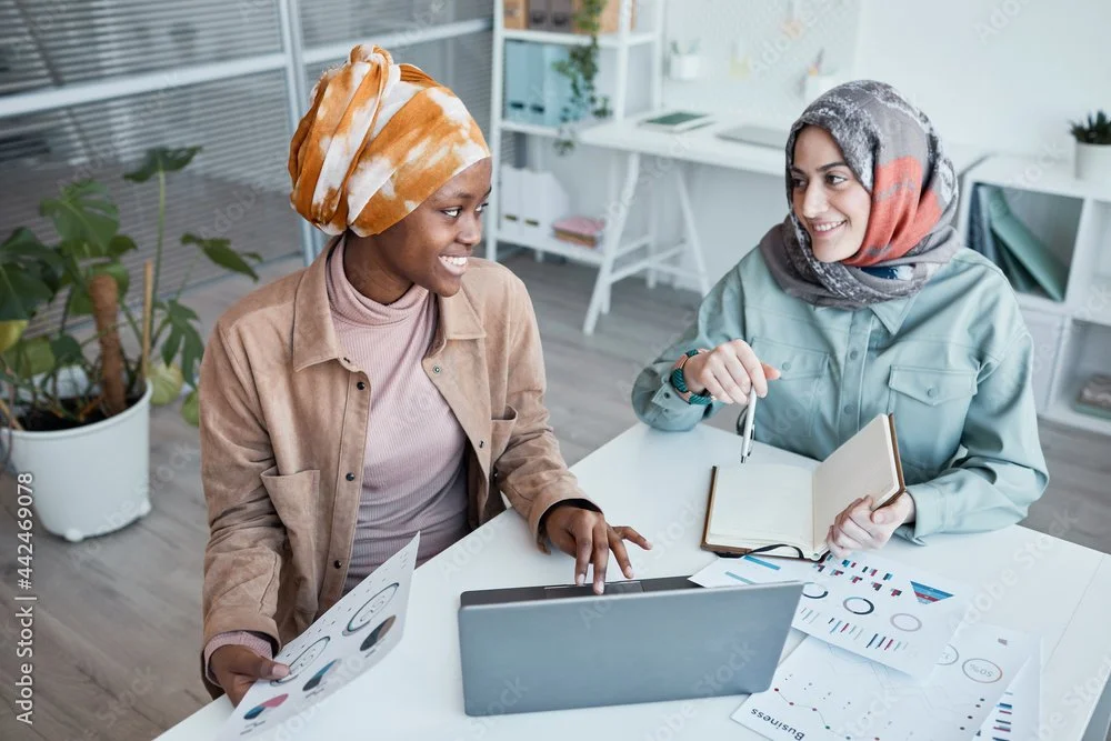 Two women, wearing headscarves, are sitting at a white desk in a modern office, smiling and engaging in a discussion while looking at each other. They have various documents, charts, and a laptop in front of them.