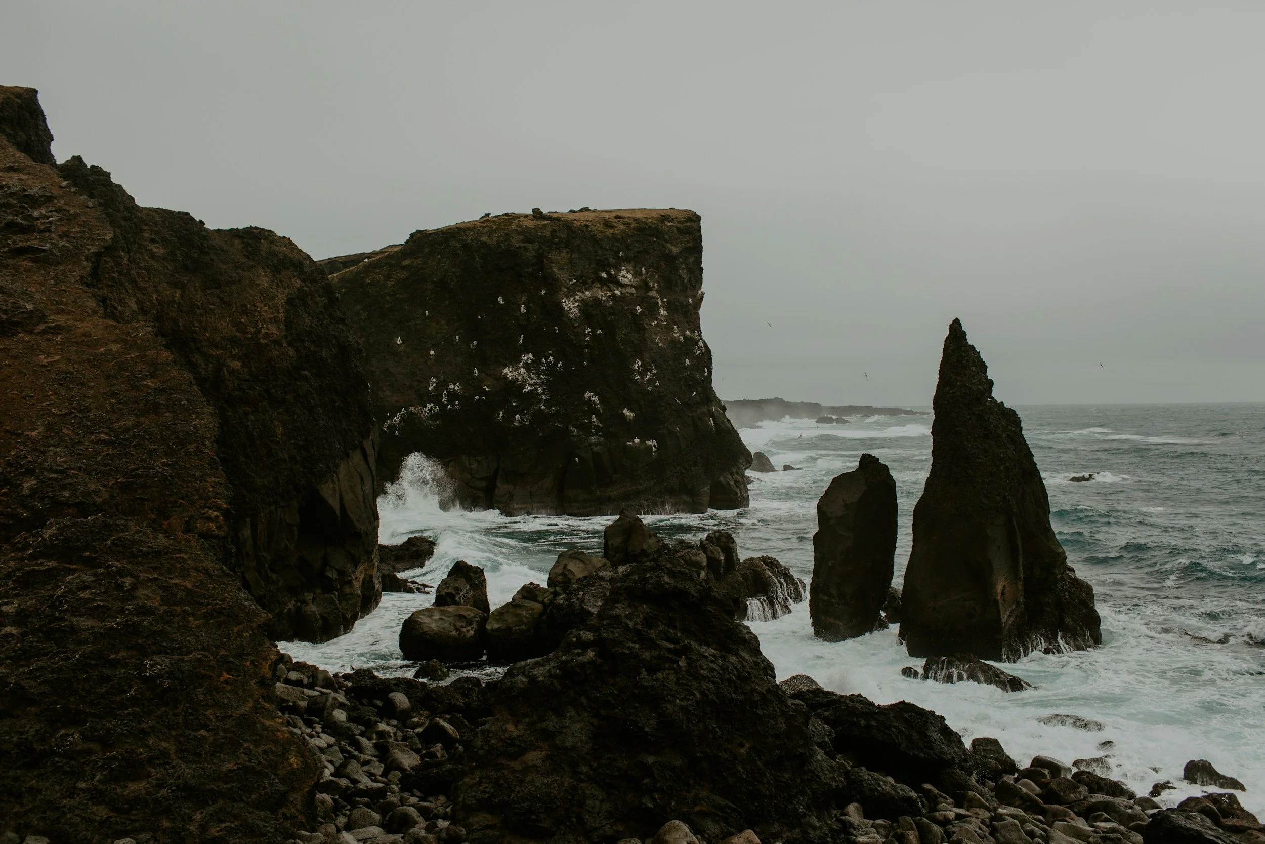 Rocky coastline with large cliffs and sea stacks in rough ocean water under an overcast sky.