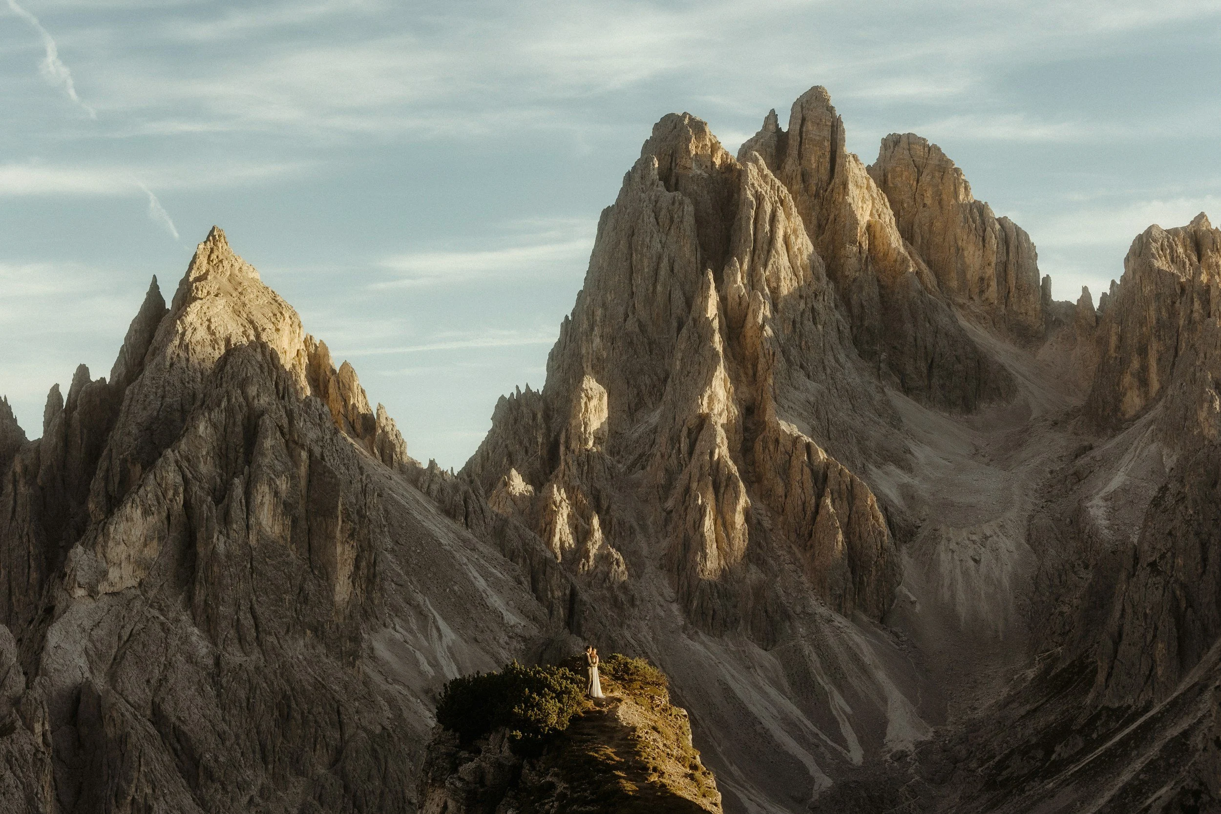 A person standing on a small hill with green bushes, overlooking rugged mountain peaks under a cloudy sky.