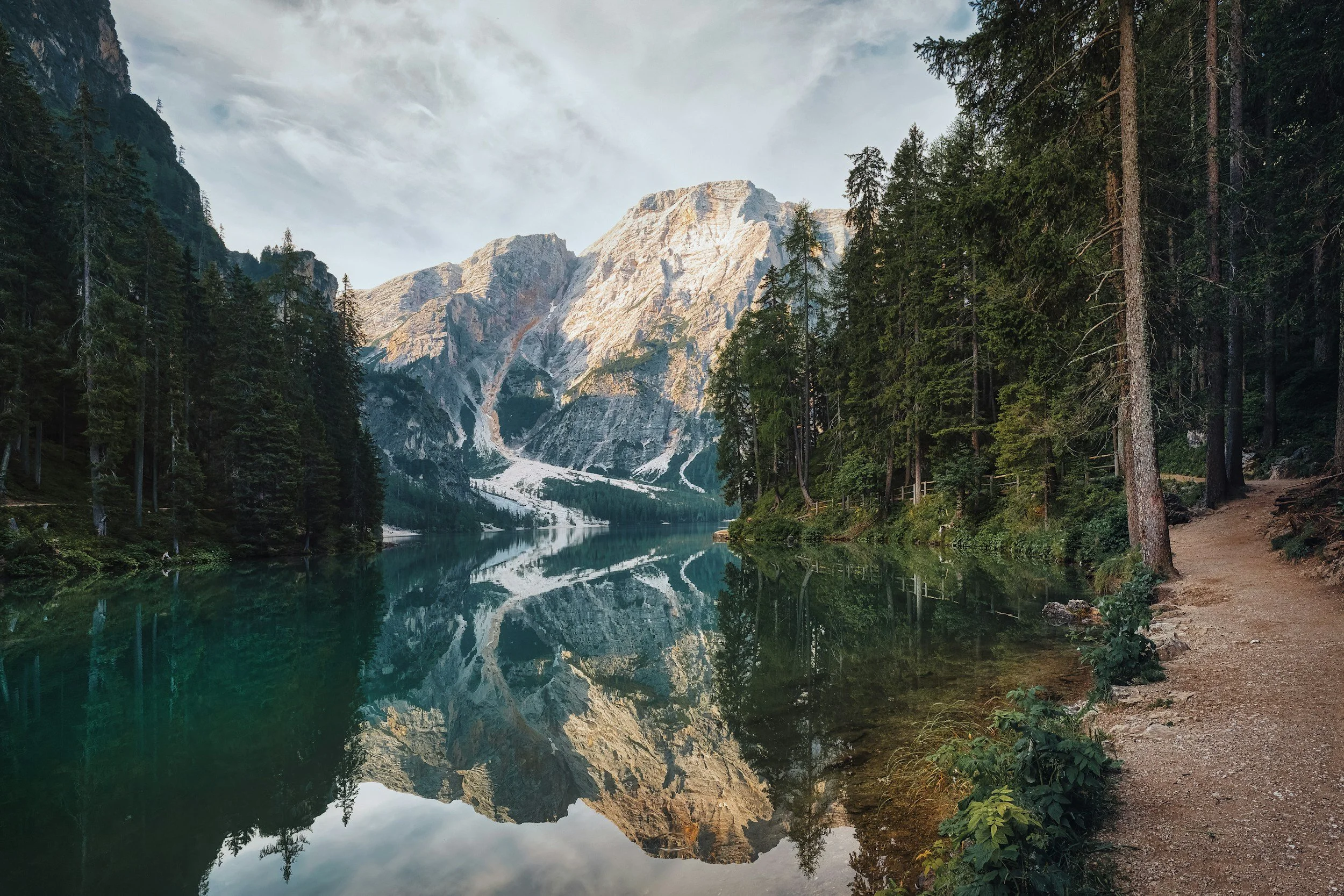 Mountain landscape with a calm lake reflecting the mountains and trees, and a dirt trail on the right side