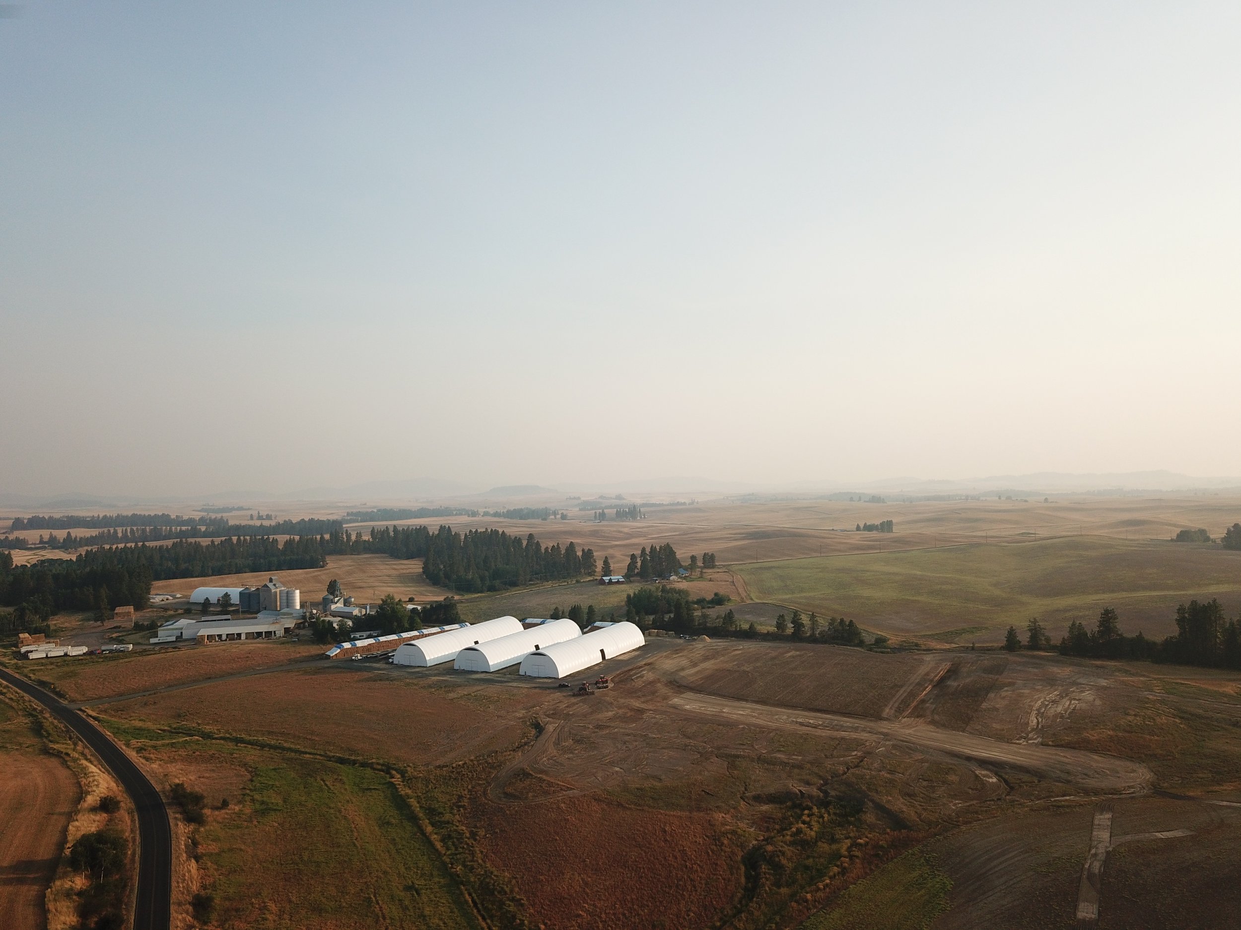 A rural landscape with farm buildings, green fields, and a winding road under a hazy sky.