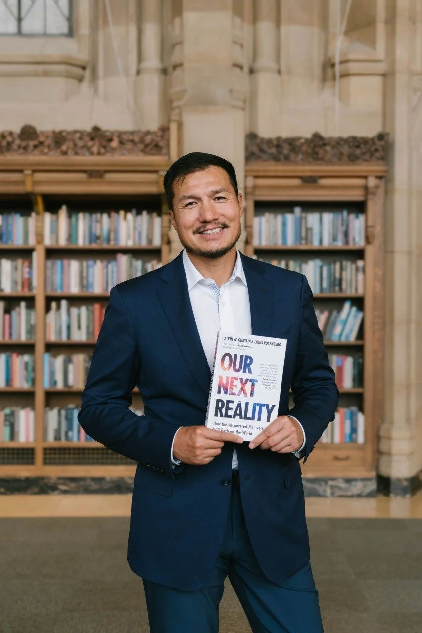 Man in a navy blue suit holding a book titled 'Our Next Reality' in front of wooden bookshelves filled with books in a library.