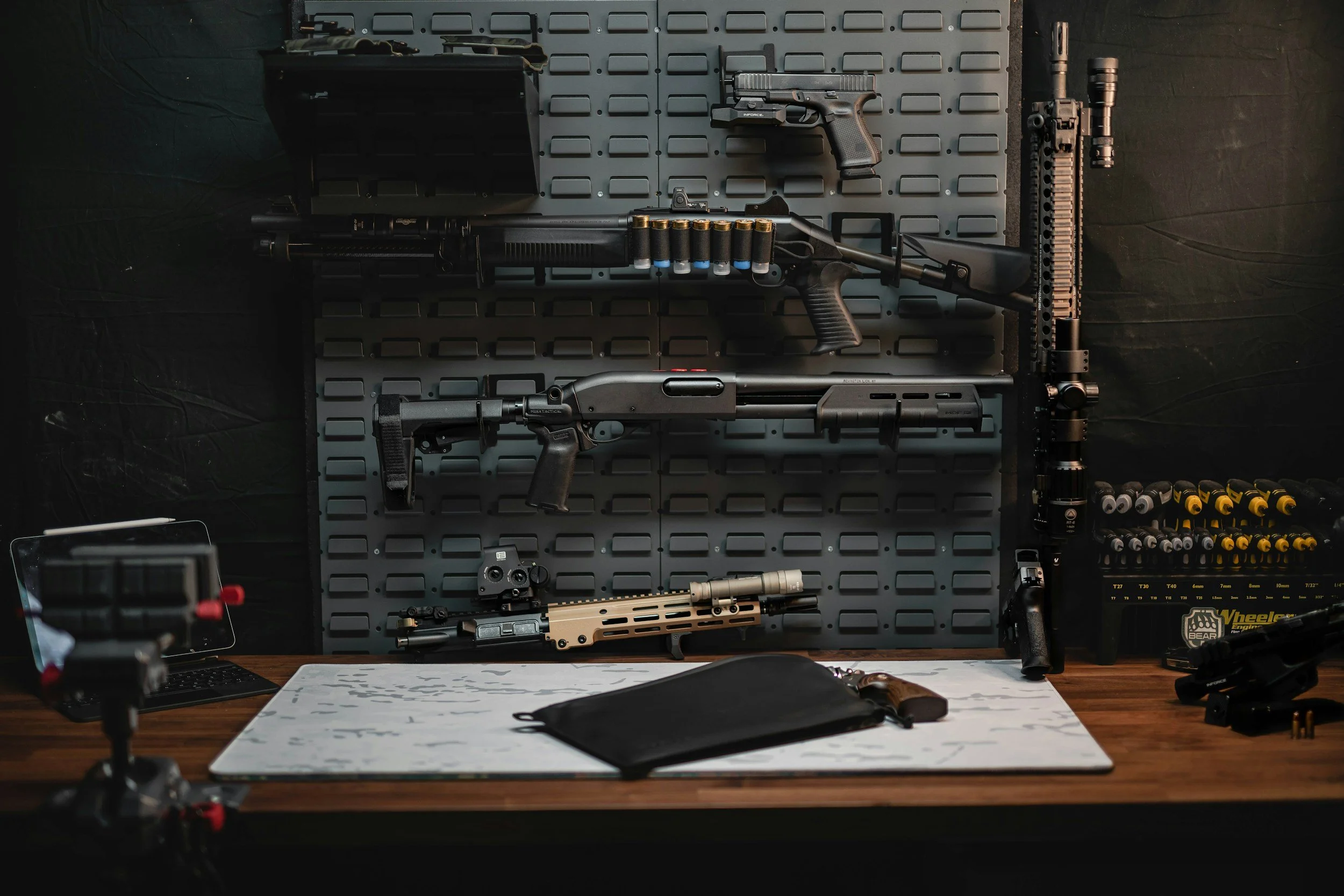 A firearms and shooting accessories setup on a workbench, including a large black rifle with a scope, a pistol, a shotgun with a tactical stock, a box of ammunition, a range bag, and a small tripod camera in the foreground. The background has a black pegboard with various attachments and a collection of screwdriver bits.