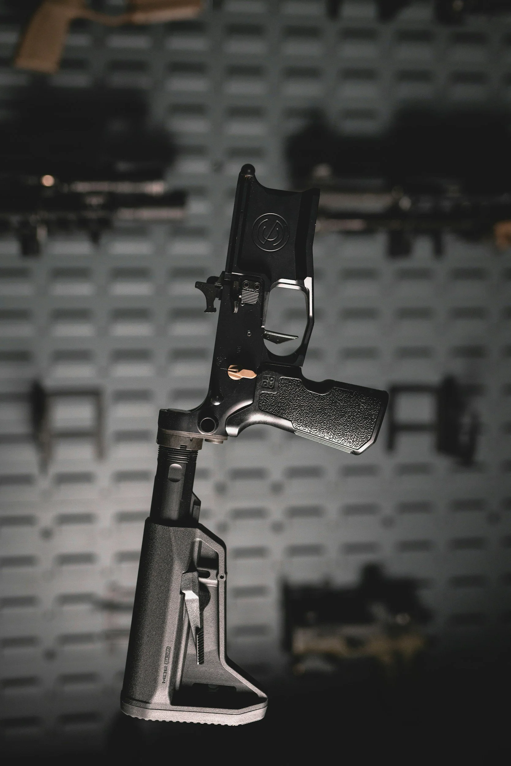 A firearm with a black slide and a textured grip is mounted vertically against a blurred background of a gray pegboard with tools and parts.