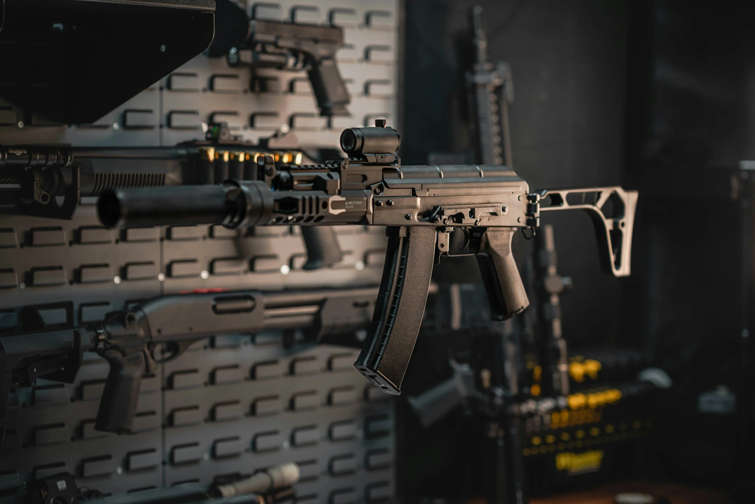 A modern, black semi-automatic rifle mounted on a display wall in a gun store or shooting range, with other firearms visible in the background.