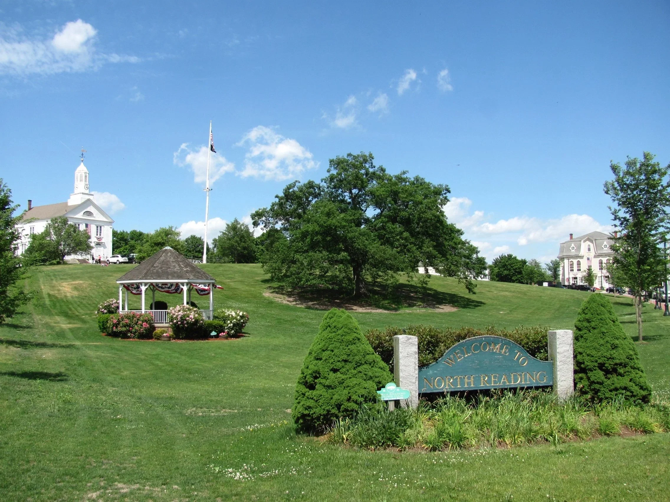 A sunny day in North Reading, with a well-maintained grassy park area, trees, a gazebo decorated for a celebration, and white houses in the background beneath a blue sky with scattered clouds.