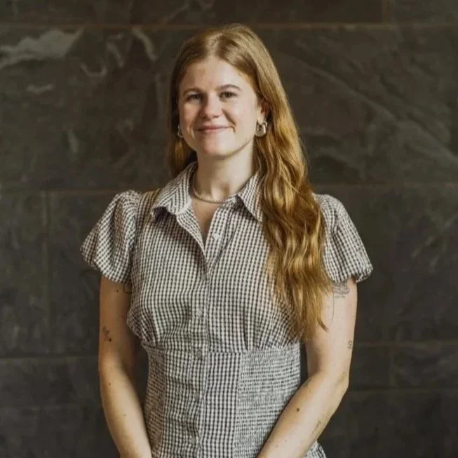 Photograph of Lucy Vehslage – A woman with long, wavy red hair, wearing a checked short-sleeve shirt, standing in front of a dark textured wall, smiling at the camera.