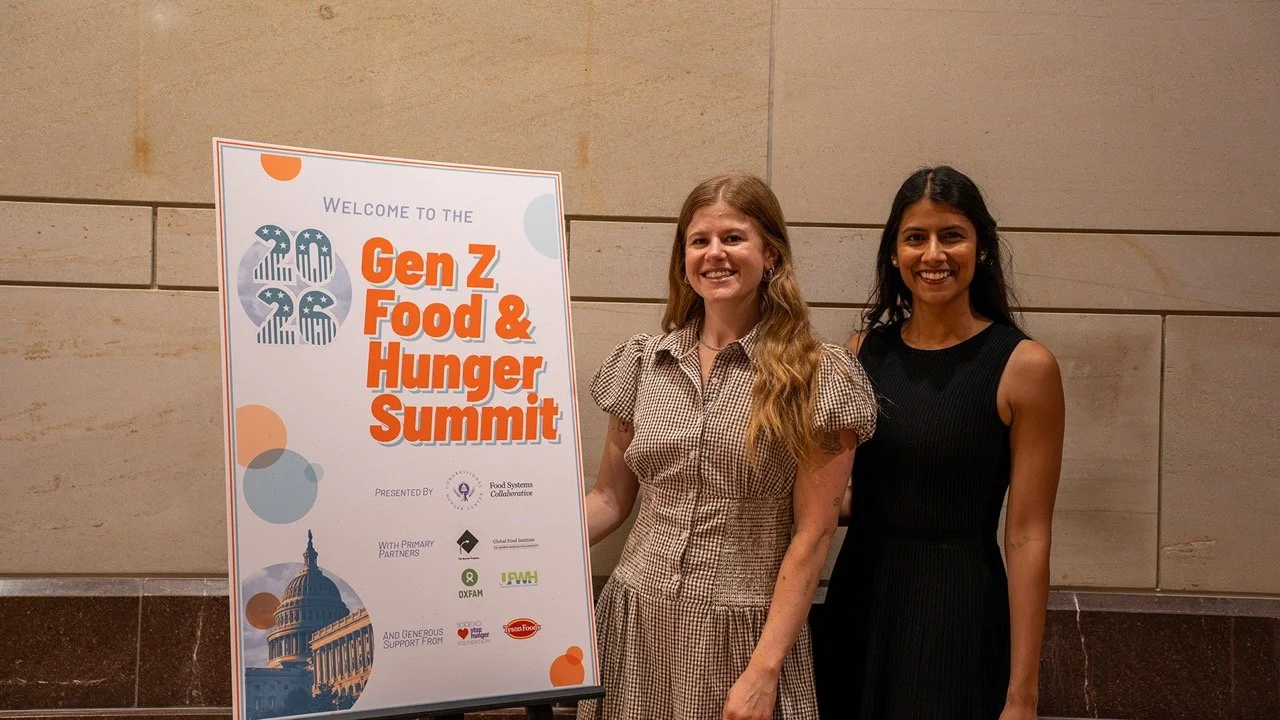Lucy Vehslage (left) and Niyeti Shah (right) stand next to a sign for the Gen Z Food & Hunger Summit, smiling at the camera.