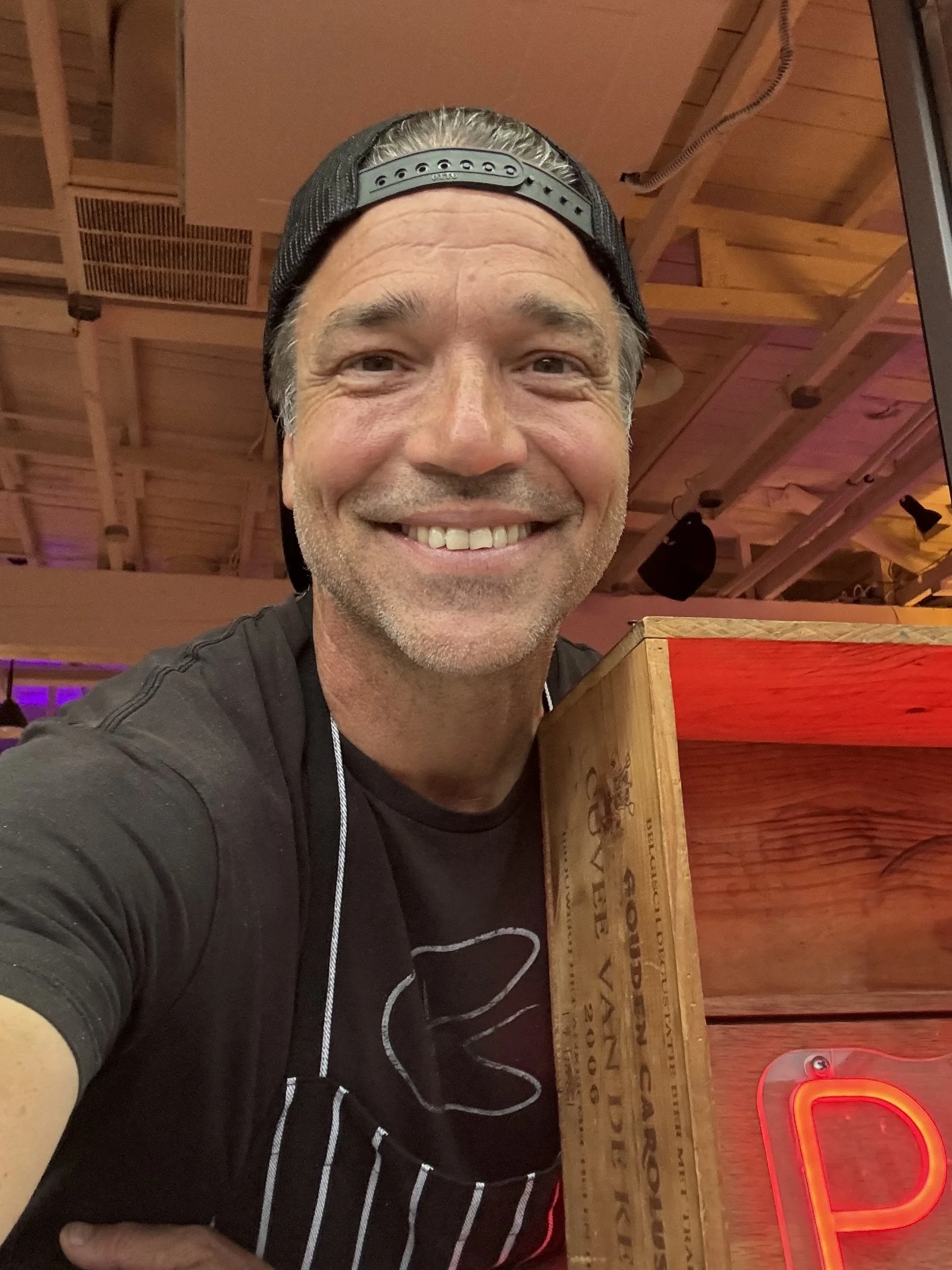 A smiling middle-aged man wearing a black cap back and a black T-shirt with white design. He is in a restaurant with wooden ceilings, a neon red pizza sign and wooden decor.