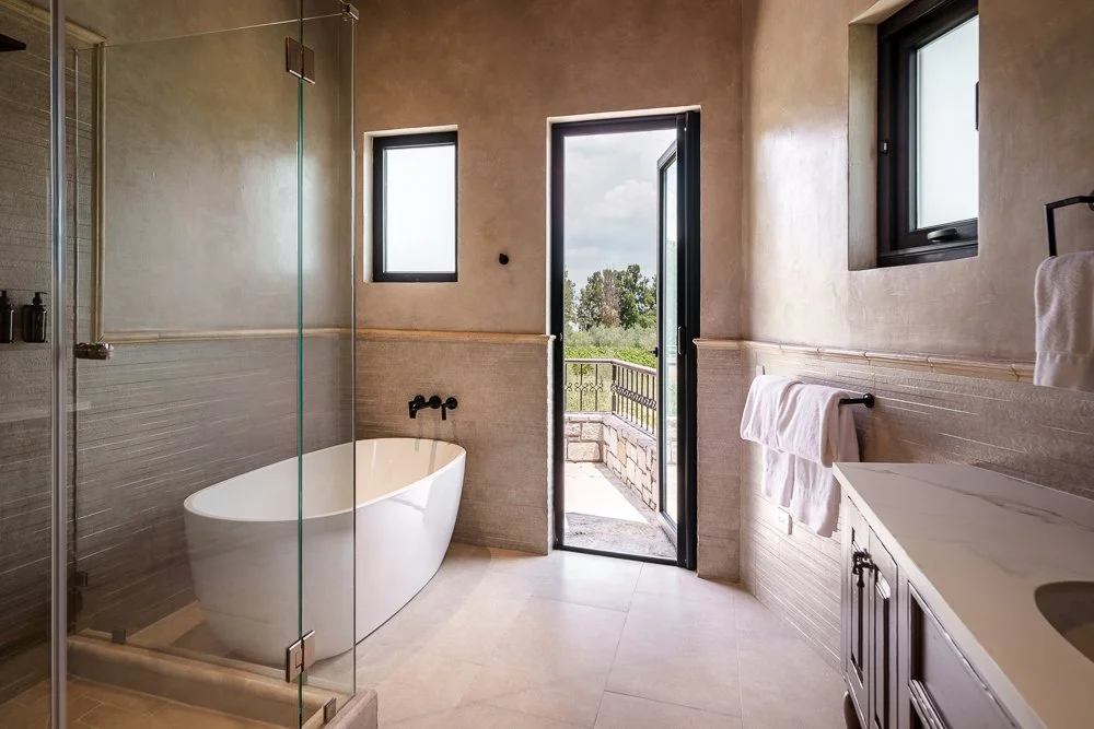 Modern bathroom with a freestanding bathtub near two small windows and a glass door leading to a balcony. There is a vanity with a marble top and towel rack, and a walk-in shower enclosed with glass. San Miguel de Allende luxury real estate photograp