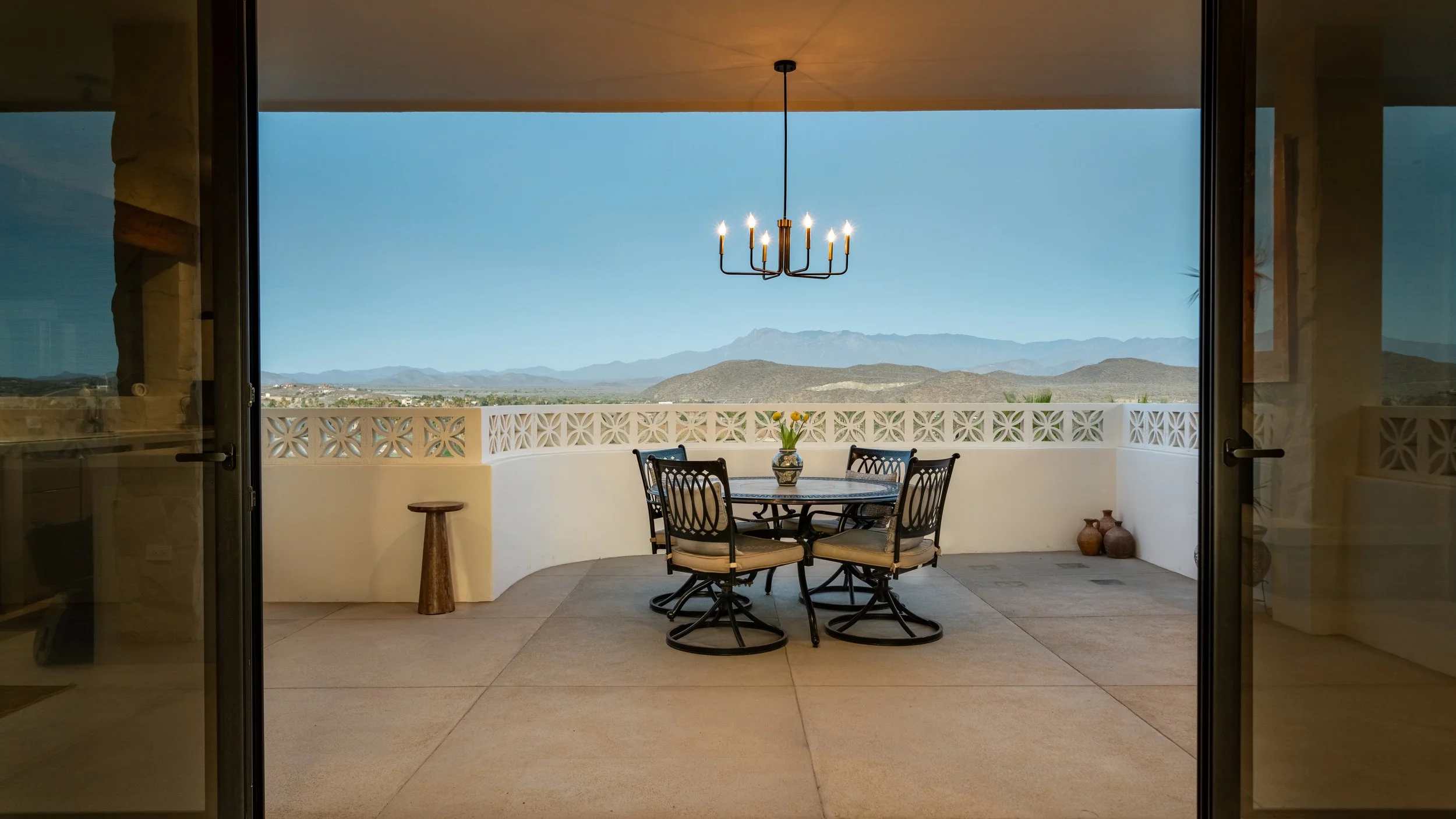 View of a balcony with a round dining table and four chairs, a vase with yellow flowers on the table, a small wooden stool, and decorative pots on the floor, with mountains in the background under a blue sky.