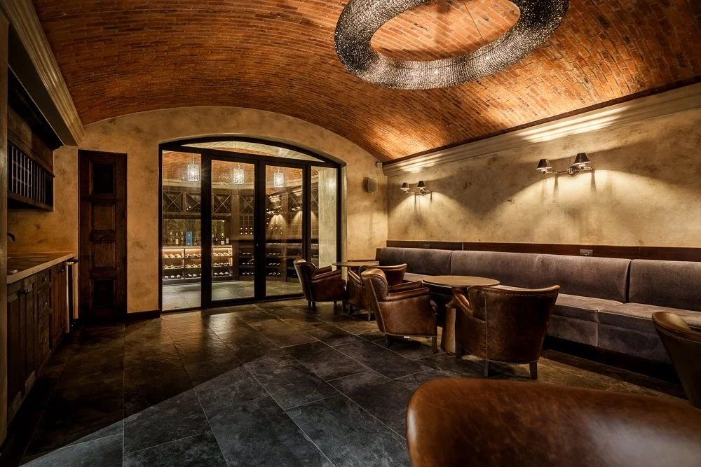 Interior of a cozy wine cellar lounge with leather chairs, a long gray cushioned bench, stone flooring, warm wall lighting, and a large glass door revealing wine bottles inside. San Miguel de Allende luxury real estate photography.