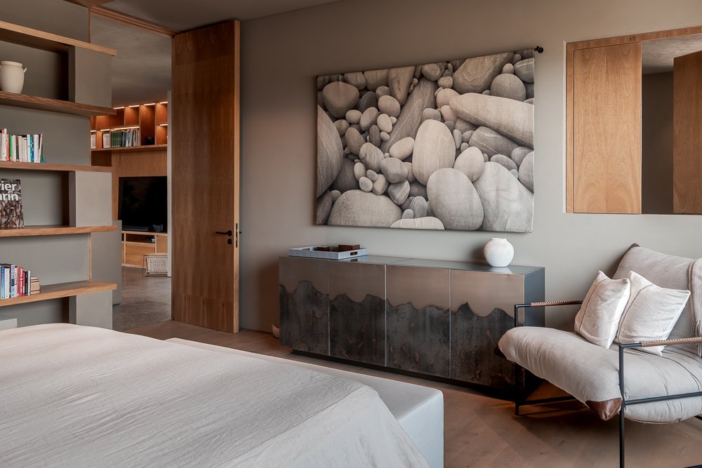 A modern bedroom with a bed, a bookshelf, and a black and gray sideboard. There is a large wall art print of white and gray stones, and a beige armchair with pillows. San Miguel de Allende luxury real estate photography.