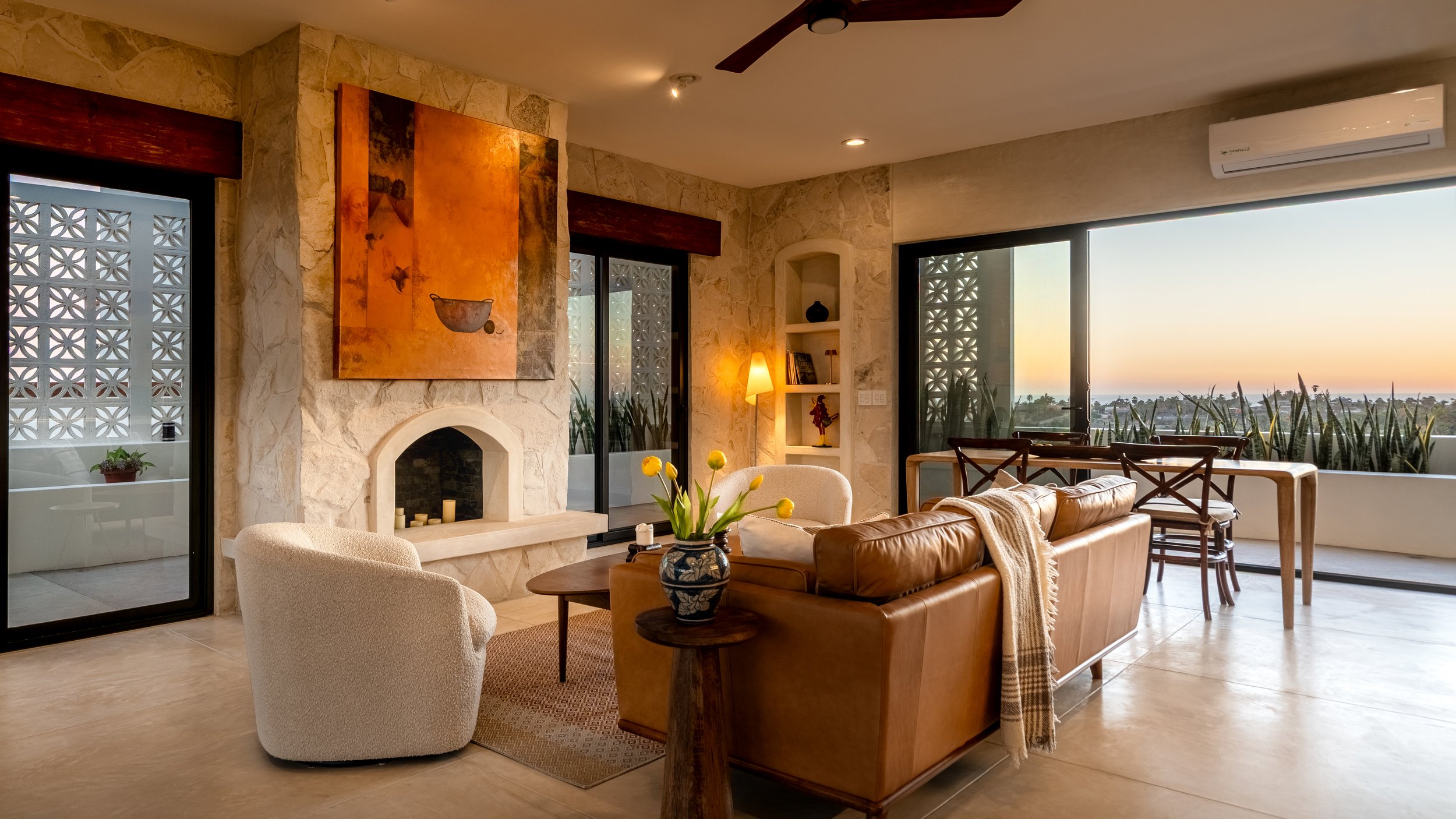 Living room with stone fireplace, large windows showing a sunset view, tan leather sofa, white armchair, wooden dining table with chairs, and a vase of yellow flowers.
